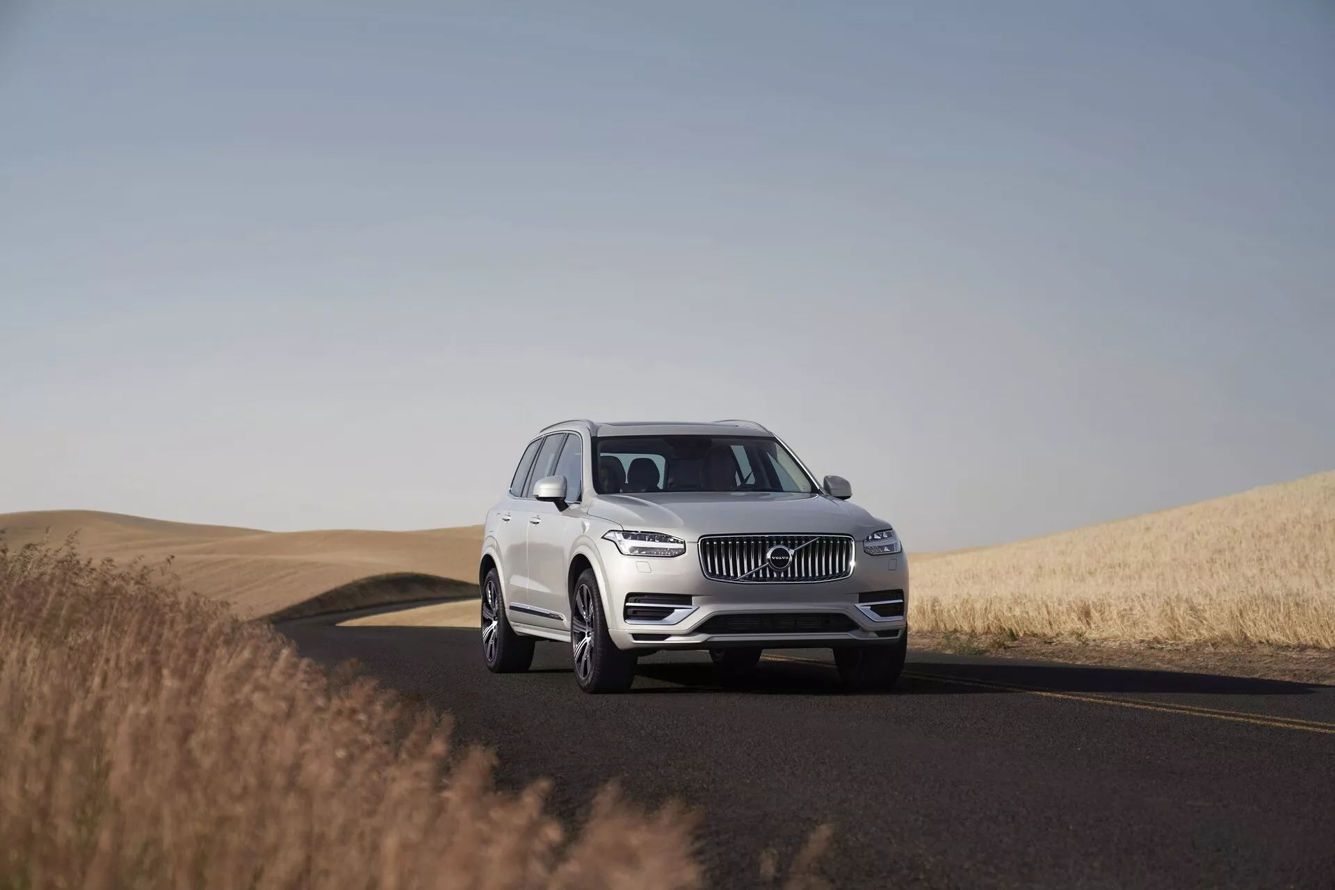 Bílé SUV Volvo XC90 z roku 2015 na dvoupruhové silnici, zachycené za denního světla s jasným nebem. Obrázek ukazuje vozidlo z nízkého až středního úhlu, což zvýrazňuje jeho impozantní přítomnost a rozměry. Pozicováno ve středu rámu s tříčtvrtečním pohledem, fotografie zachycuje detaily designu jako mřížku, LED světlomety, emblém a přední nárazník, což naznačuje pokročilé asistenční systémy řidiče. Kola, slitiny a případně velikost pneumatik jsou jasně viditelné. Klidné prostředí obsahuje silnici zahýbající doprava a zlatá pšeničná pole, což zdůrazňuje vhodnost vozidla pro dlouhé cesty v různorodé krajině.