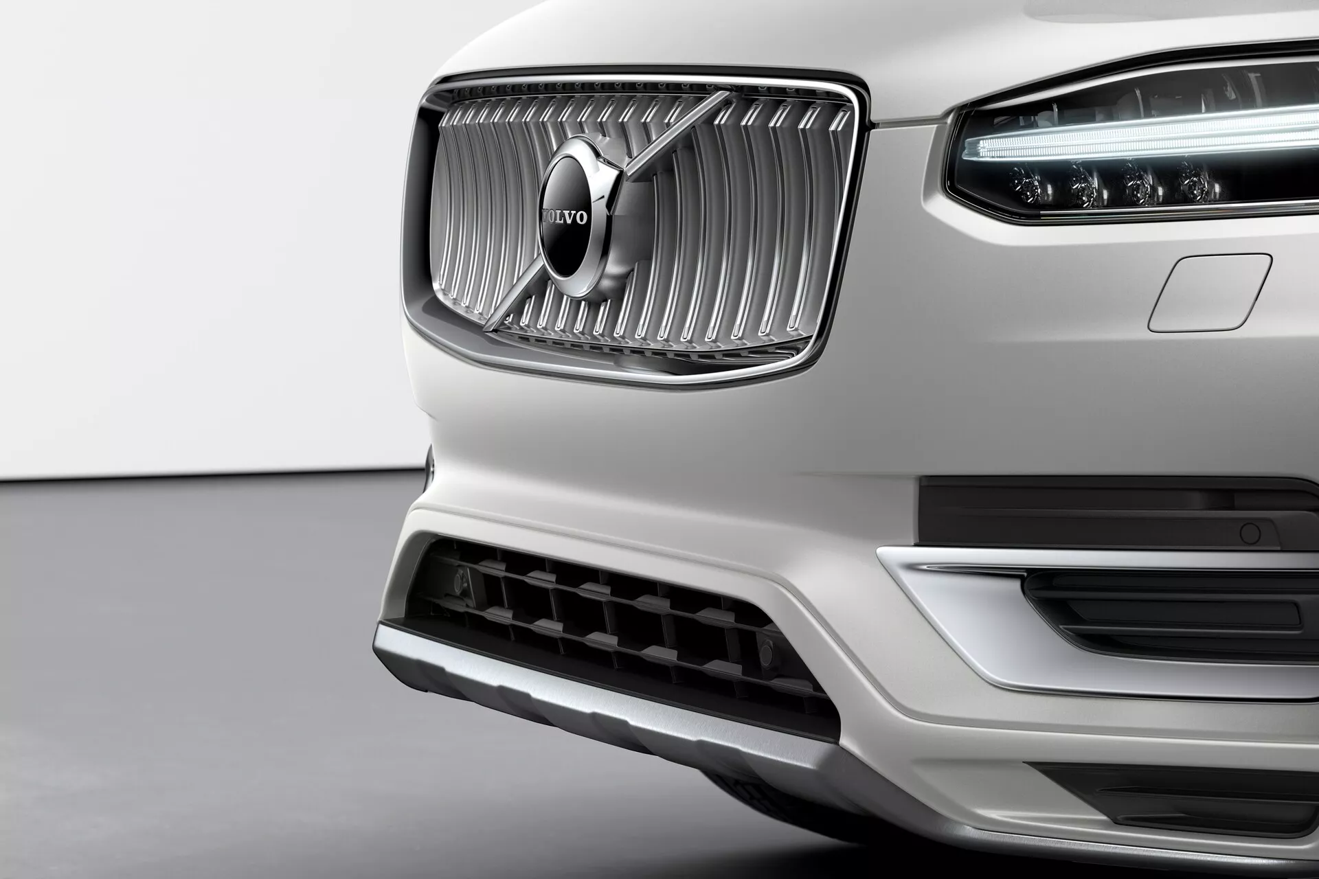 Detailní záběr přední části bílé Volvo XC90 2015 druhé generace s výraznou mřížkou s vertikálními lamelami a charakteristickým znakem Volvo uprostřed. Chromový povrch mřížky kontrastuje s matnou texturou kolem ní. Viditelné jsou LED denní světla Thor's Hammer v hlavním světlometu. Spodní nárazník obsahuje velký přívod vzduchu s ozdobným stříbrným lemem, což naznačuje aerodynamickou účinnost a chlazení motoru. Záběr z mírně nižšího úhlu zdůrazňuje eleganci, kvalitu zpracování a prémiový přední design značky Volvo.