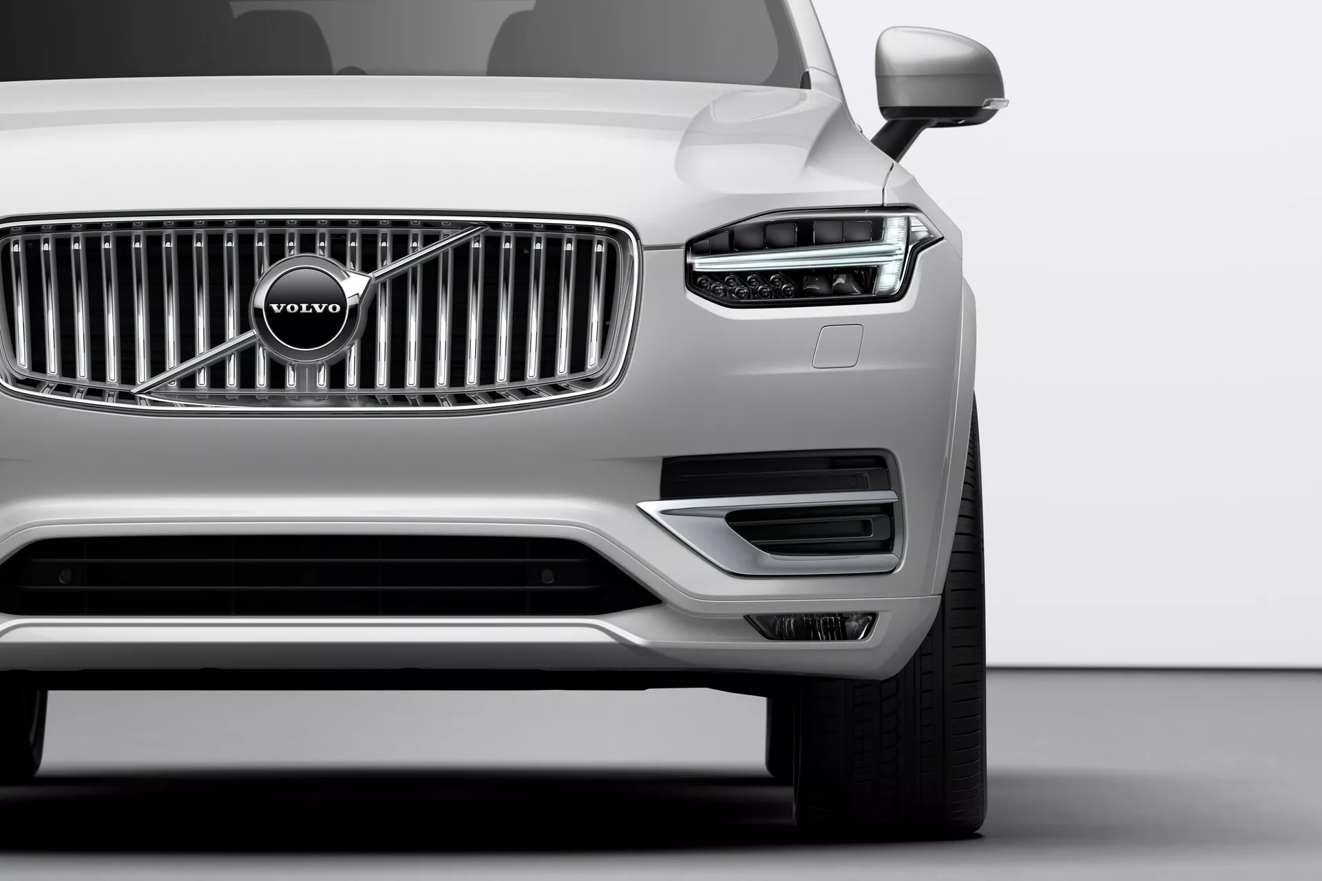 Detailní záběr přední části stříbrného vozu Volvo XC90 z roku 2015 (2. generace) pořízený z nízkého úhlu. Zdůrazněny jsou zejména chromované mřížky s ikonickým znakem Volvo, elegantní světlomety s denními LED světly ve tvaru T nazývanými 'Thorovo kladivo' a dobře integrované parkovací senzory na předním nárazníku. Dolní část nárazníku obsahuje nasávací otvory a kryty mlhových světel zdobené stříbrným lemováním. Viditelná zrcátka naznačují pokročilé technologické prvky jako ukazatele směru nebo varovné systémy pro mrtvý úhel.