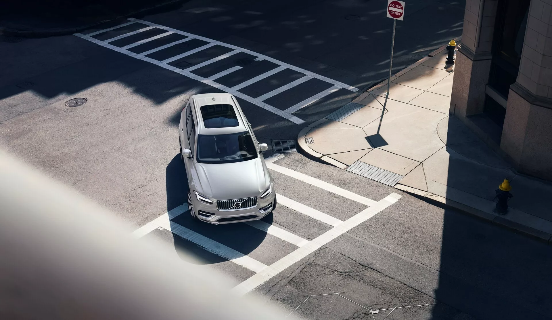 Snímek shora vozu Volvo XC90 z roku 2015 (2. generace) z vysokého úhlu, diagonálně z předního levého rohu; zobrazující světlou kapotu vozu, střechu a část strany řidiče. Vůz je správně zaparkován na vyznačeném místě u přechodu. Světlomety naznačují aerodynamické tvary a moderní designové linie. Ve městě zvýrazňují situaci dopravní značky a stíny naznačující polední osvětlení. Vůz také naznačuje moderní technologické funkce bez viditelných senzorů nebo kamer.