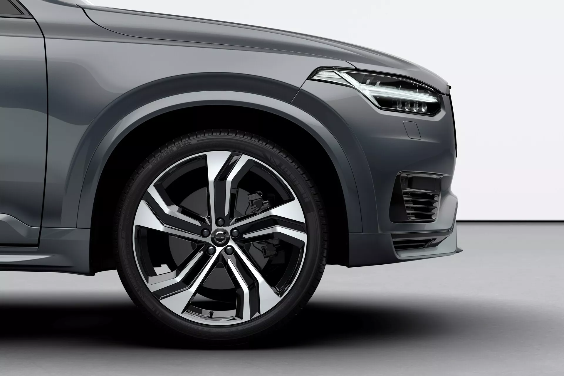 Nízký úhel snímku předního kola na straně řidiče a přilehlých částí vozu Volvo XC90 2015. Verze ukazuje moderní hliníkový ráfek s několika paprsky, sportovní pneumatiku s nízkým profilem, část předního blatníku s aerodynamickými liniemi, elegantní design světlometu, přední nárazník s horizontálním vzorem mřížky, možný mlhový světlomet nebo větrací otvor pod světlometem a kovový lak, to vše naznačuje luxusní nebo vyšší výbavu vozidla.