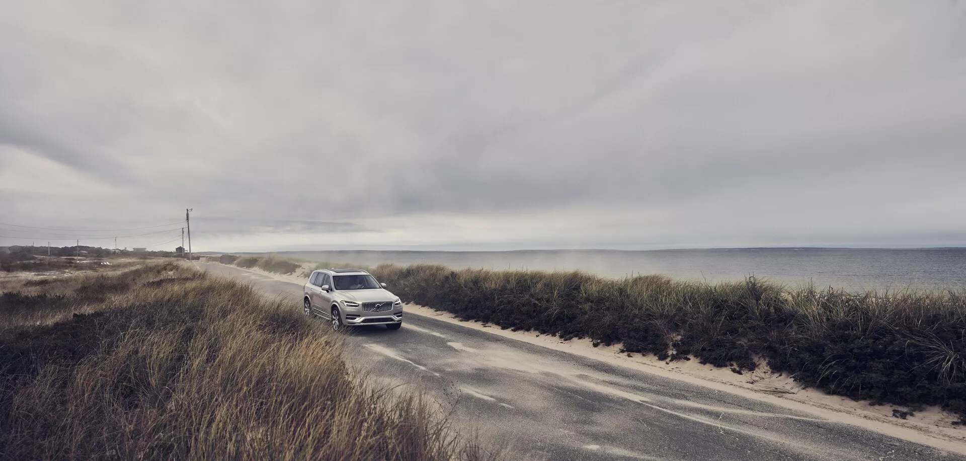 Fotografie Volvo XC90 (2015, 2. generace) jedoucího po úzké pobřežní silnici. Auto přibližující se ke kameře zdůrazňuje přední mřížku, světlomety a přední nárazník. Na pozadí zatažené oblohy a oceánu SUV s lehkou kovovou barvou vyniká. Tato scéna zdůrazňuje spojení luxusu a dobrodružství, naznačuje pokročilé technologické a bezpečnostní funkce pro pobřežní jízdní podmínky.
