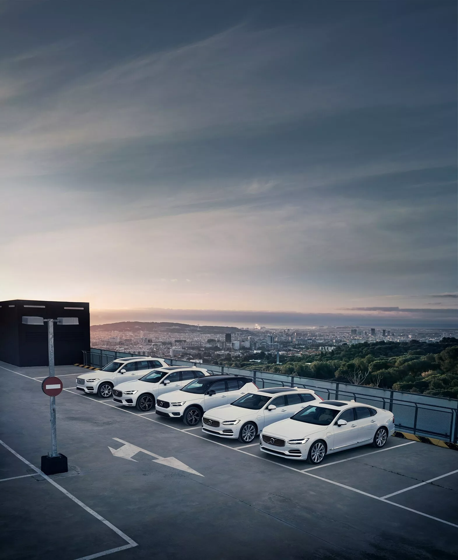 Řada čtyř moderních, luxusních vozidel Volvo XC90 2015 (2. generace) zaparkovaných na horním patře zvýšeného parkovacího objektu během východu nebo západu slunce s městskou krajinou v pozadí. Bílé sedany mají aerodynamický design, vícepaprsková hliníková kola, aerodynamická boční zrcátka a tónovaná okna, čímž předvádějí své stylové kontury a dynamickou estetiku.