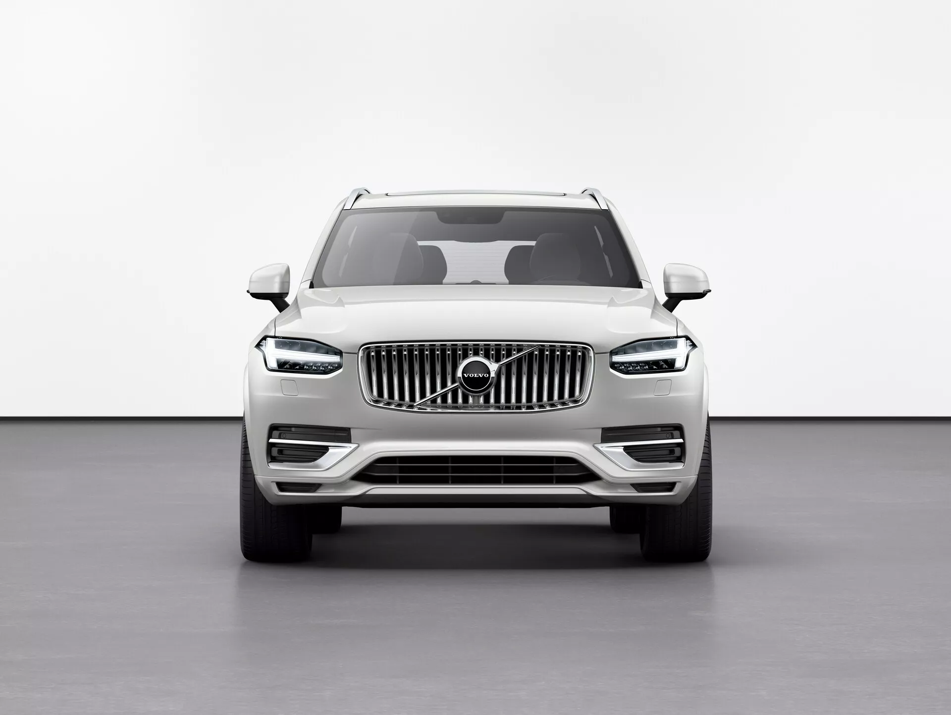 Přední pohled na vůz Volvo XC90 2015 (2. generace) SUV proti bílému pozadí ve studiovém nastavení, zdůrazňující vyšší světlou výšku, velkou mřížku s vertikálními pruhy s emblémem Volvo, elegantní LED světlomety 'Thorovo kladivo', výrazné linie kapoty, mlhová světla na spodním nárazníku a boční zrcátka s integrovanými směrovkami.