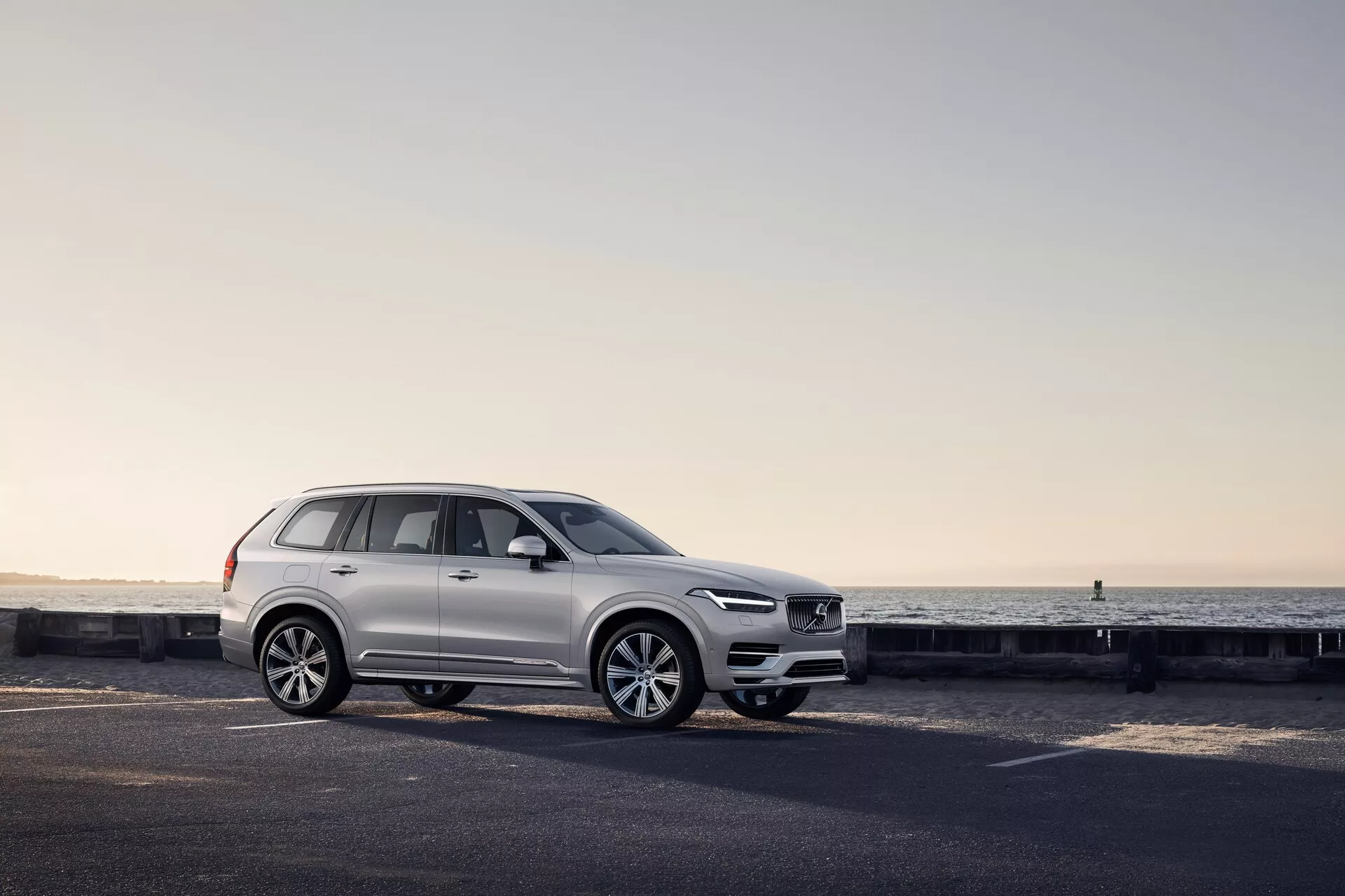 Boční profil bílého SUV Volvo XC90 2015 na pobřežní silnici s viditelnými designovými prvky, včetně designu kol, dveřních panelů, bočních zrcátek, střešních ližin a konfigurace oken zvýrazněné měkkým svítáním nebo soumrakem.