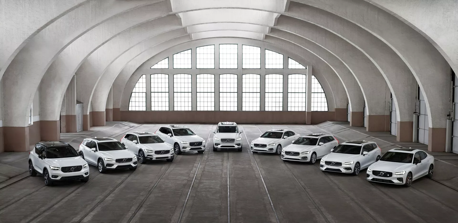 Řada osmi bílých vozů Volvo XC90 2015 druhé generace stojí uvnitř prostorné, architektonicky klasické budovy s velkými klenutými okny a klenutým stropem. Vozidla jsou natočena směrem ke kameře v různých úhlech a předvádí své přední části a designové prvky, včetně LED světel, velkých slitinových kol a tvarovaných karoserií. Osvětlení zdůrazňuje designové prvky aut, zatímco podlaha, označená stopami pneumatik, naznačuje, že prostor je využíván k parkování vozidel.