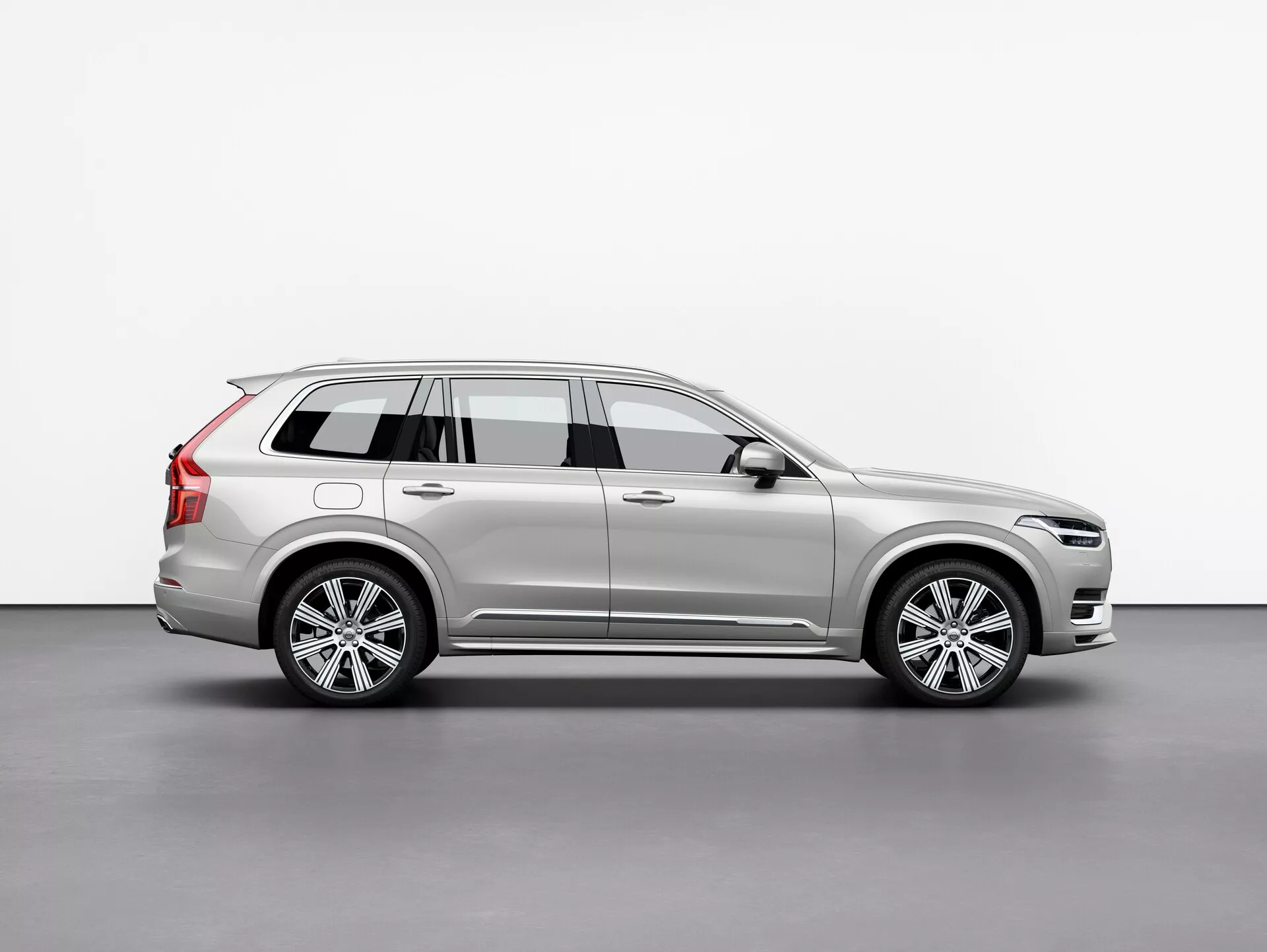 Boční profil stříbrného vozu Volvo XC90 2015 (2. generace) SUV, zobrazující jeho hliníková kola, střešní nosiče, boční zrcátko s integrovaným směrovým světlem a detailní linie karoserie, umístěný centrálně proti jednoduchému pozadí.