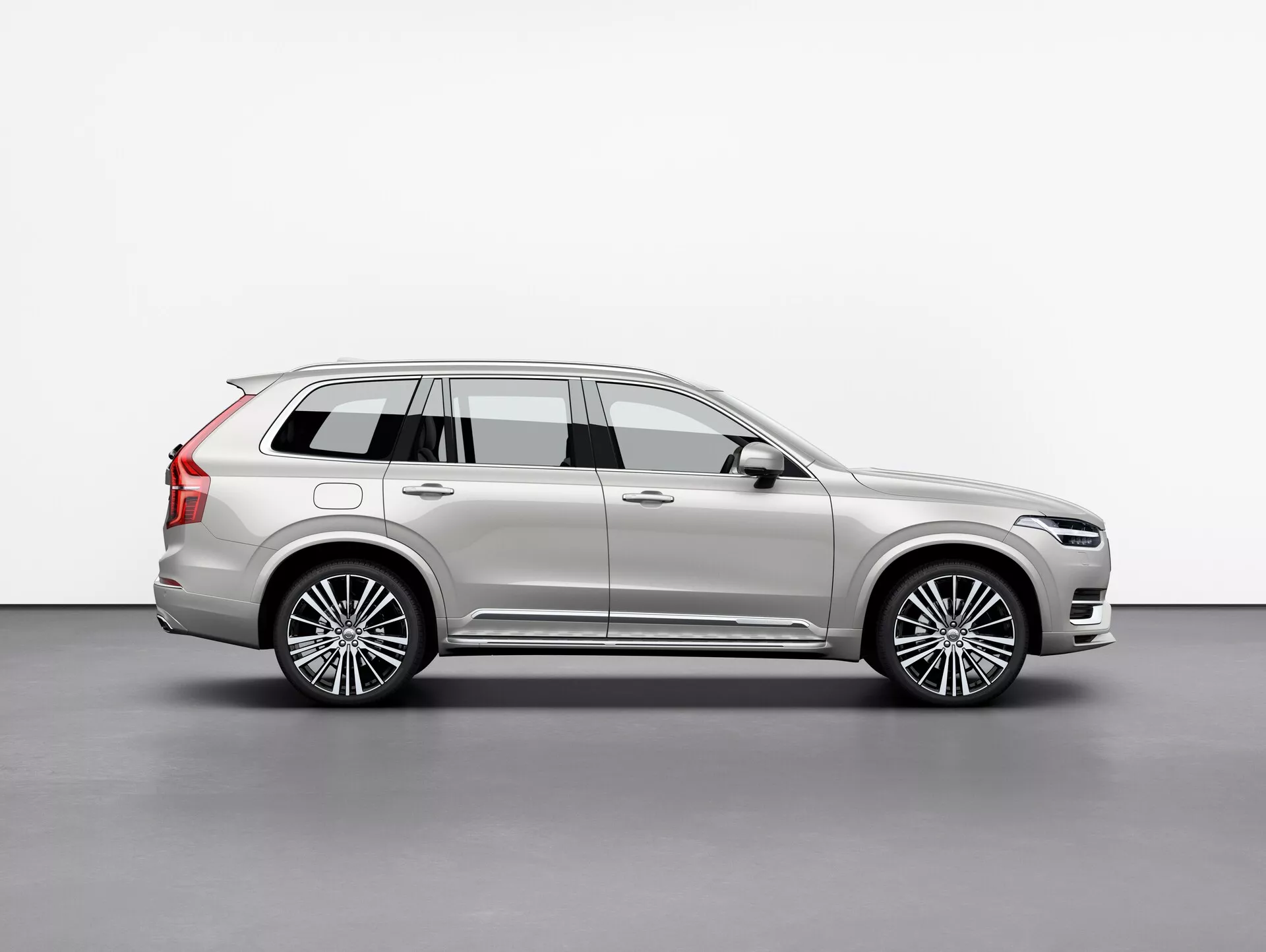Boční profil vozu Volvo XC90 2015, luxusního SUV plné velikosti, zachycený ve studiu na bílém pozadí. Obrázek ukazuje dlouhý rozvor, čtyři dveře, zadní výklopnou stěnu, vícepaprsková litá kola, chromované lišty, metalický lak a výrazné designové prvky jako boční zrcátka a střešní lišty. Pečlivě osvětlená scéna zvýrazňuje linky a křivky karoserie, které odrážejí její prémiovou povahu. Vozidlo je umístěno tak, aby poskytovalo jasný pohled na celkovou délku, výšku a postoj, což svědčí o SUV s kapacitou tří řad pro cestující a náklad.
