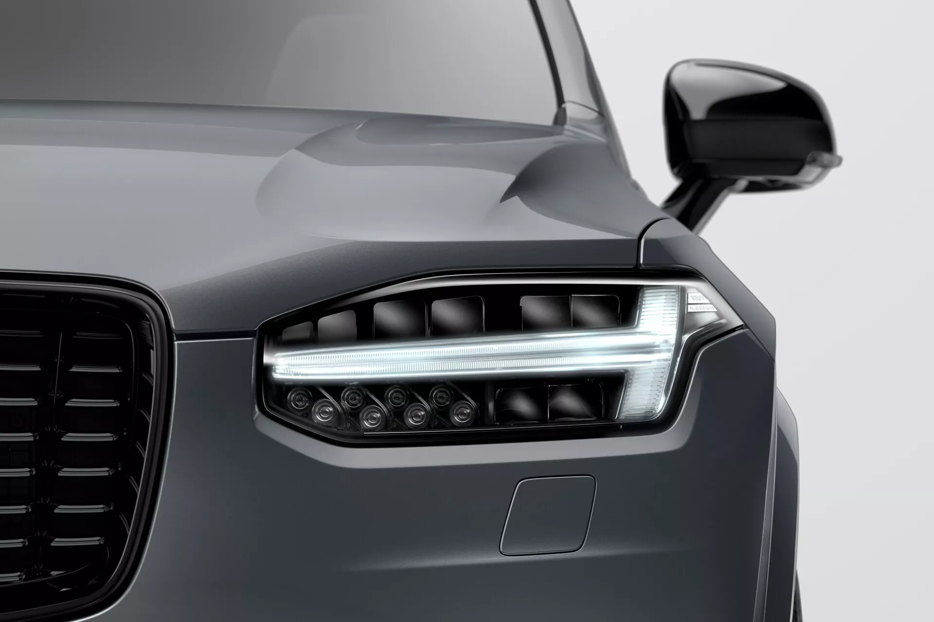 Detailní záběr na přední část Volvo XC90 2015 (2. generace), zaměřený na levý světlomet s moderní LED nebo laserovou technologií, dvojitými osvětlovacími prvky, elegantními denními běžícími světly a několika projektorovými čočkami pro lepší viditelnost. Obrázek zdůrazňuje velkou esteticky navrženou masku chladiče, aerodynamické tělo zrcadla v barvě karoserie a možné ADAS čidlo pod světlometem. Lesklý, odrazivý povrch tmavé metalické šedě zdůrazňuje elegantní, avšak svalové linie kapoty a blatníku, což ukazuje na vysoce kvalitní lakování a špičkový design vozidla.