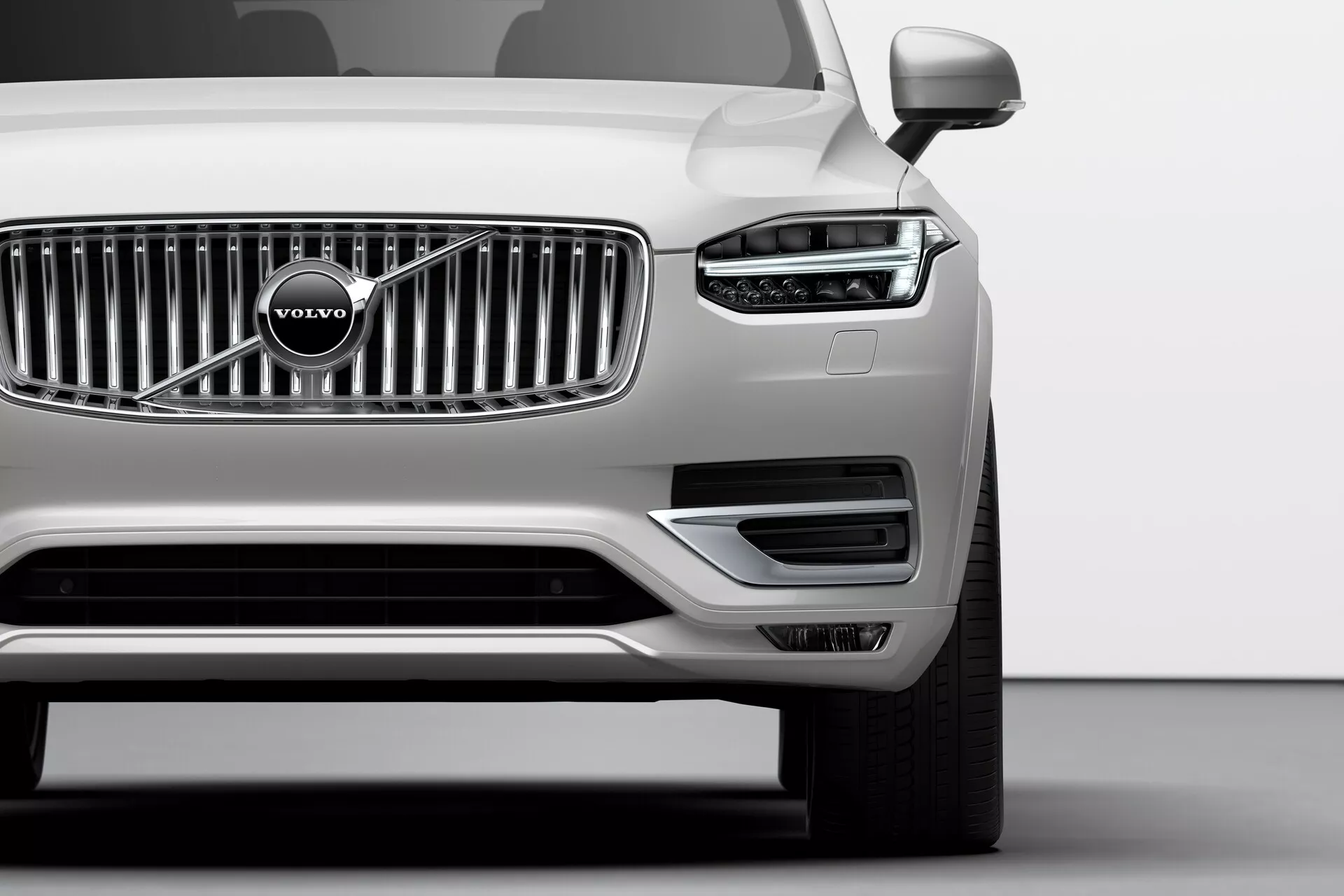 Nízký úhel stříbrného Volva XC90 (2. generace) z roku 2015 s pohledem na přední levý čtvrtinový díl v ateliéru, s charakteristickým LED světlem Thorovo kladivo, vertikálně chromovanou mřížkou chladiče s emblémem Volvo, minimalistickým předním nárazníkem s nasáváním vzduchu, levým zrcátkem, vícepaprskovým slitinovým kolem a bezchybným metalickým lakem, které ilustrují pokročilou světelnou technologii, design značky a aerodynamické prvky.