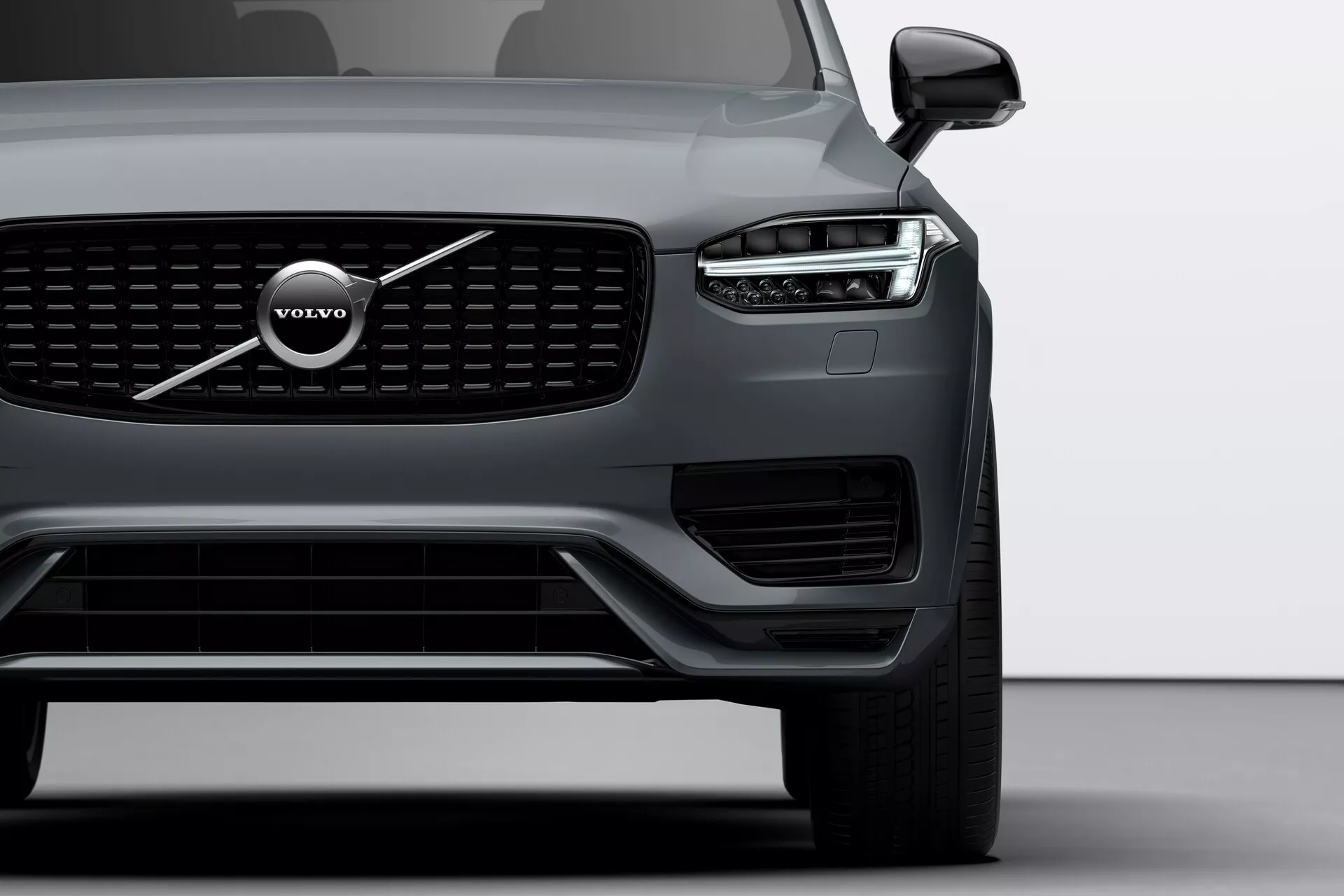 Detailní záběr z předního úhlu na Volvo XC90 (druhá generace, 2015) zaměřený na levou stranu, s výraznou mřížkou, velkým znakem Volvo a LED světlometem 'Thorovo kladivo'. Obrázek zahrnuje světlomet, přední kolo, část dveří řidiče, viditelné senzory na spodním nárazníku a zrcátko s možnou technologií sledování slepého úhlu.