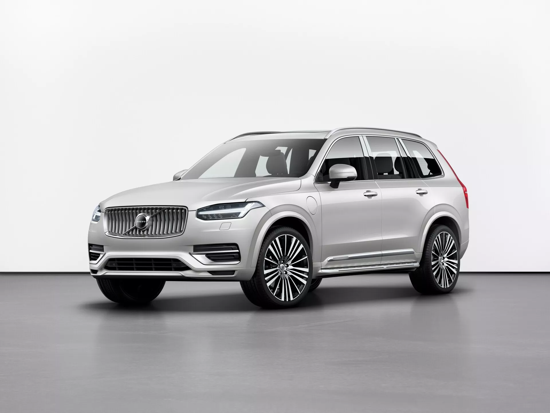 Ateliérová fotografie luxusního SUV Volvo XC90 2015 (2. generace) zobrazeného z úhlu tří čtvrtin zepředu, která ukazuje přední mřížku s výrazným logem, elegantní LED světlomety, masivní přední nárazník a dlouhý rozvor s velkými litými koly. Zrcátka jsou umístěna pod sloupkem A, s okny zdobenými chromovanými lištami a střešními nosiči. Neutrální barva laku vyzařuje sofistikovanost a snímek zdůrazňuje linie a kontury vozu v rovnoměrném osvětlení proti jednoduchému pozadí. Pravděpodobné funkce zahrnují asistenční technologie řidiče a pokročilé informační systémy.