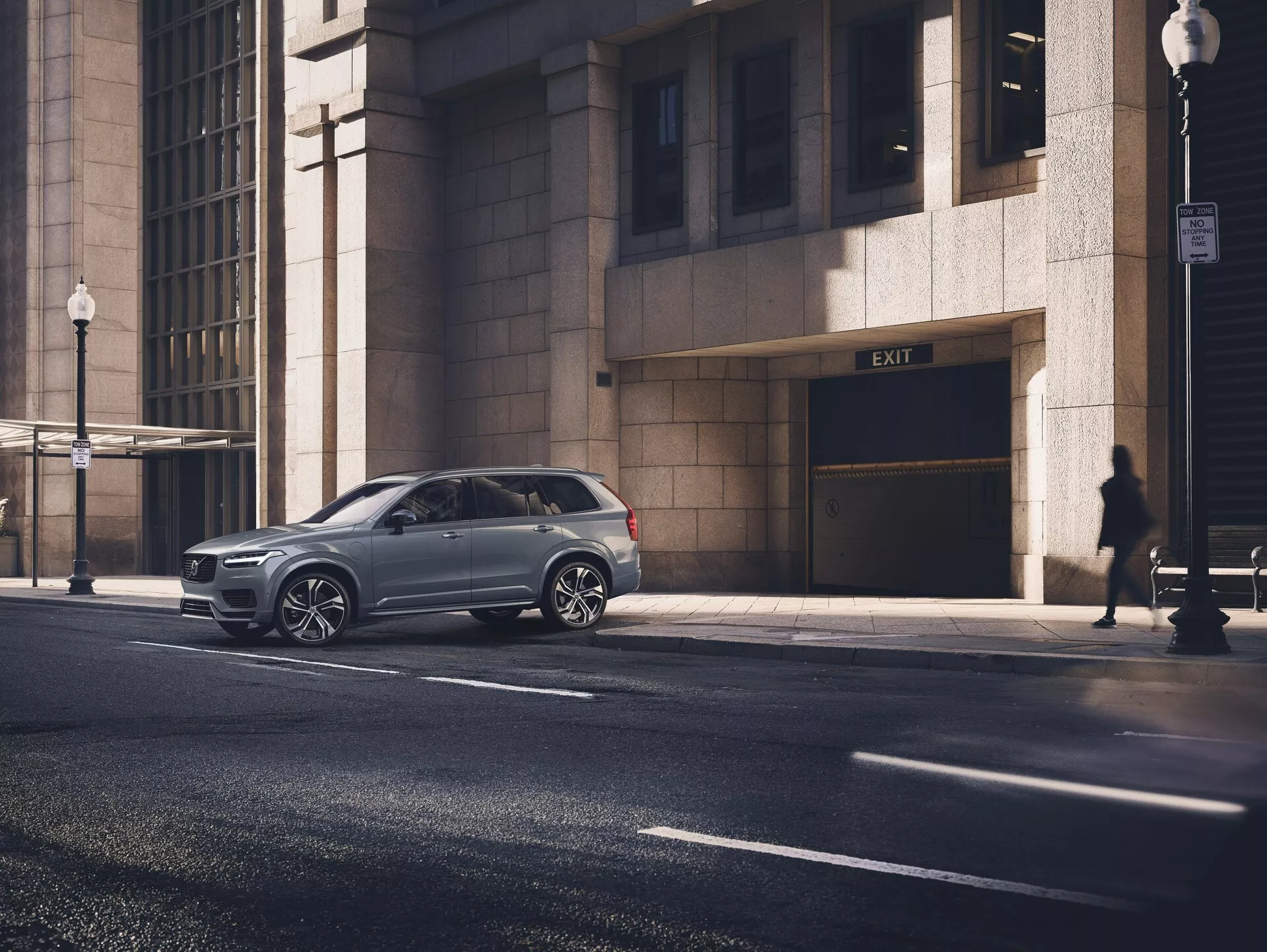 Moderní Volvo XC90 2015 (2. generace) SUV zaparkované na městské ulici, pořízené z perspektivního úhlu ukazujícího boční profil a část přední masky. Lesklý kovový lak vozu zdůrazňuje jeho dobře udržovaný stav, vícepaprsková slitá kola a tónovaná okna. Obrázek je pořízen z oční úrovně průměrné dospělé osoby, ukazuje vůz ve středu obrazu obklopený městskými prvky, včetně fasády budovy, siluety chodce a pouliční lampy za ranního nebo pozdně odpoledního slunečního světla.