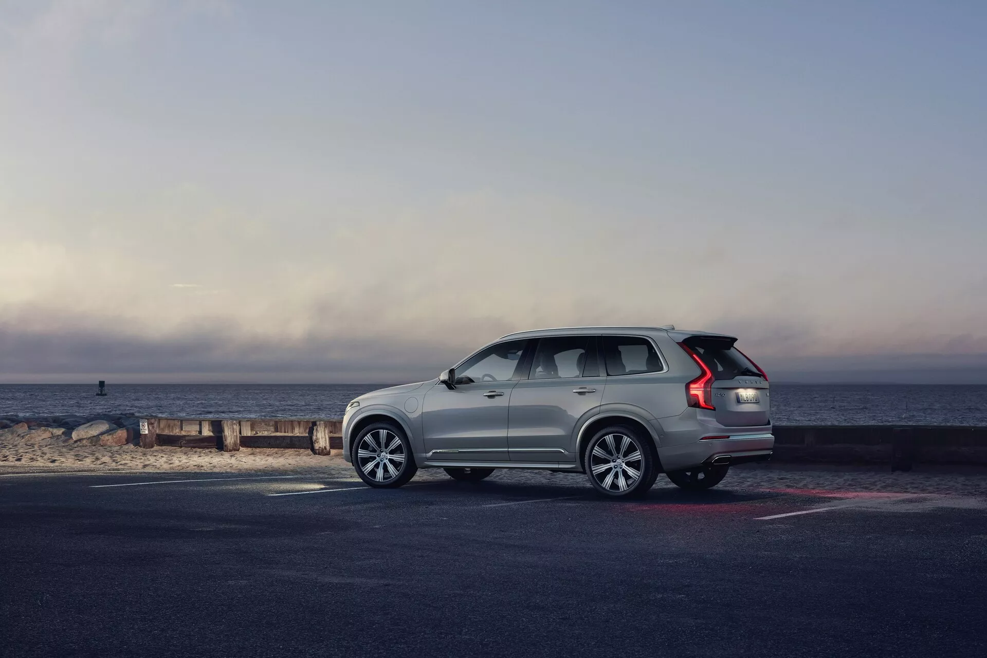 Boční pohled na SUV Volvo XC90 z roku 2015 (2. generace) zaparkované u břehu, vozidlo směřuje doprava. Úhel zdůrazňuje stranu a zadek, předvádí designové prvky, jako jsou vícepaprsková kola z lehké slitiny, prodloužená zadní světla LED, střešní ližiny, stoupající okenní linie a zapuštěné kliky dveří. Světle zbarvené SUV vyniká proti tmavšímu chodníku a pláži, osvětlené přirozeným ranním nebo pozdním odpoledním světlem, se scénickým pozadím horizontu, kde se obloha setkává s mořem. Celková kompozice vyvolává pocit dobrodružství, luxusu a svobody.