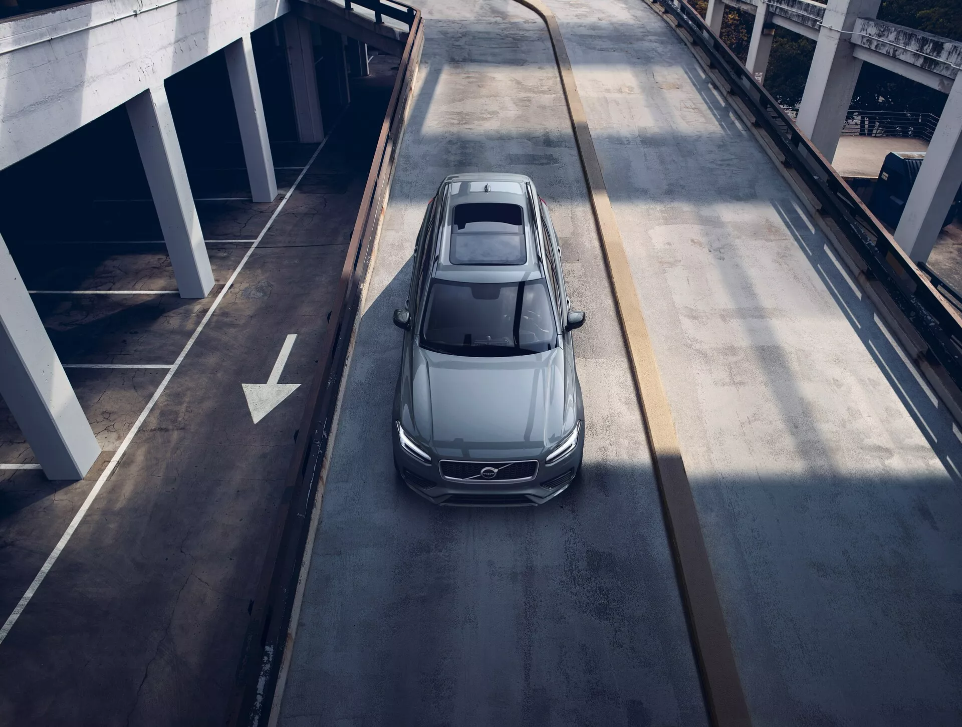 Pohled z ptačí perspektivy na šedý nebo stříbrný sedan Volvo XC90 z roku 2015, jedoucí po víceúrovňové městské silnici, ukazující panoramatickou střechu, aerodynamický design, elegantní LED světlomety a velká hliníková kola, což naznačuje moderní luxusní vozidlo.