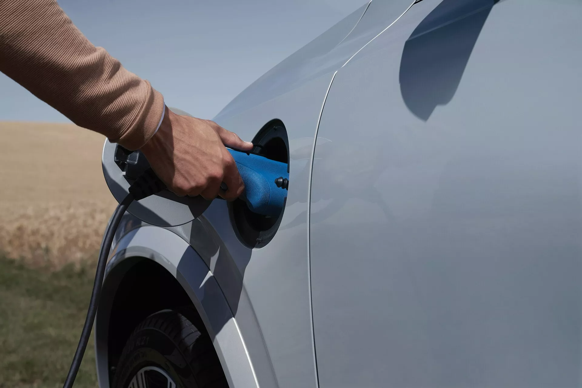 Pohled z bočního úhlu na elektromobil nebo plug-in hybridní vozidlo Volvo XC90 2015 druhé generace během nabíjecího procesu. Snímek zachycuje bok vozidla se zaměřením na nabíjecí port a konektor, částečně ukazuje zadní kolo a část spodku vozu. Modrý nabíjecí kabel je zasunut, pozadí otevřené přírodní oblasti, naznačující venkovské prostředí.