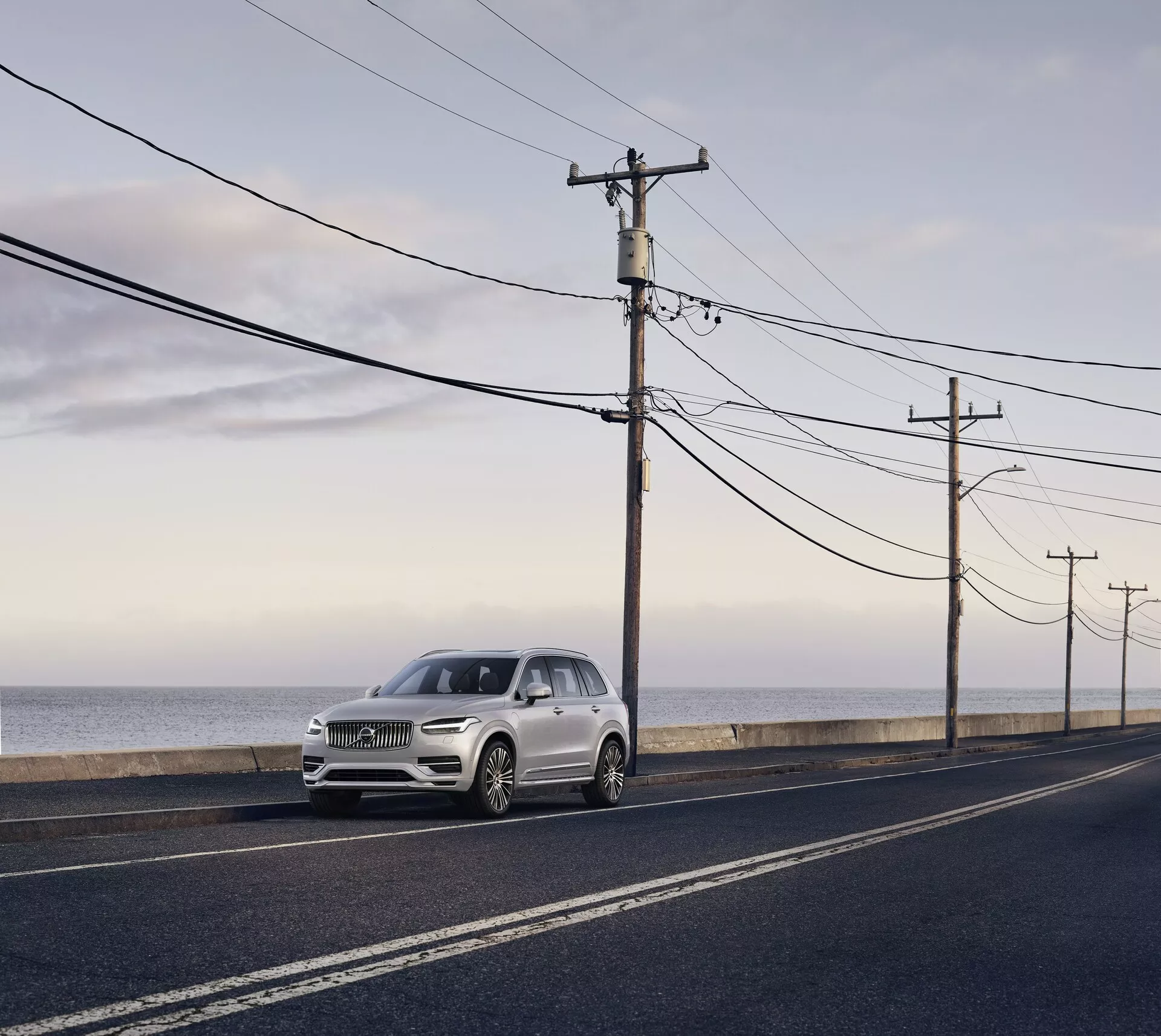 Volvo XC90 druhé generace z roku 2015, luxusní SUV zaparkované na asfaltové silnici rovnoběžné s pobřežím oceánu, pořízené ve dne s měkkým rozptýleným světlem a dlouhými stíny. Pohled z předního úhlu, zdůrazňující elegantní designové prvky včetně světlometů, mřížky a kontur kapoty, světle stříbrné nebo bledě šampaňské barvy kontrastující s tmavou silnicí a přírodním pozadím.