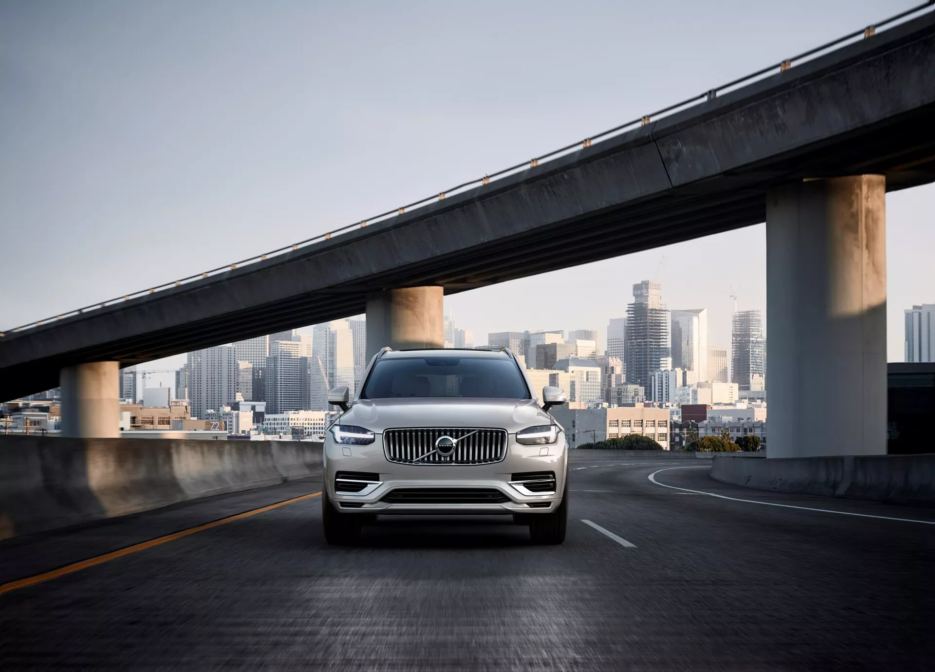 Čelní snímek SUV druhé generace Volvo XC90 z roku 2015 na prázdném mostě s panorámou města v pozadí. Úhel kamery je nízký, zdůrazňující přední mřížku, LED světlomety, chromové akcenty, litá kola a tvarovaný nárazník se senzory, což naznačuje asistenci při parkování nebo systém prevence kolize. Perleťová nebo kovová barva vozu září v denním světle, zdůrazňující jeho luxusní a moderní design v městském prostředí.