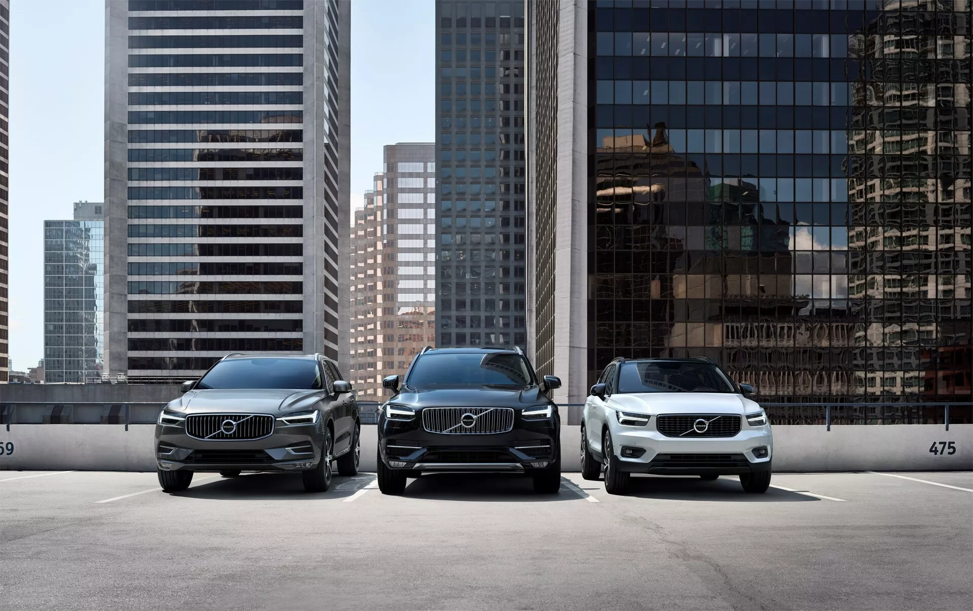 Tři luxusní vozy Volvo XC90 2015 (2. generace) zaparkované vedle sebe, čelí fotoaparátu na pozadí moderní městské krajiny. Přední perspektiva vozidel ukazuje elegantní LED světlomety, velké grily s výraznými emblémy Volvo a čistý, elegantní design. Auto v popředí na pravé straně je světlejší barvy, což zdůrazňuje různé barevné možnosti. Jsou zaparkované za obrubníkem s viditelným číslem 475, což naznačuje vyhrazenou parkovací plochu nebo výstavní prostor.
