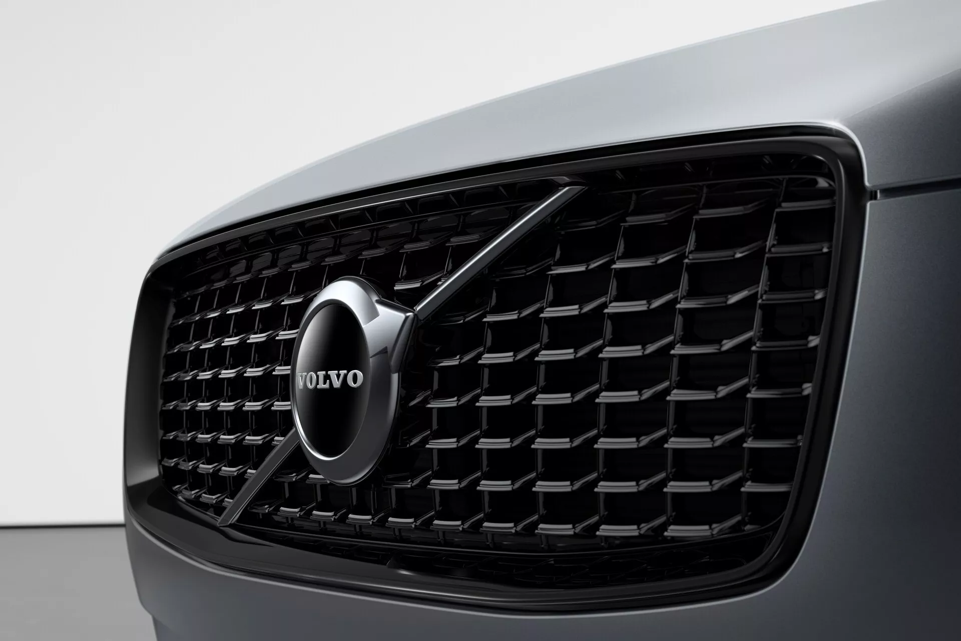 Detailní záběr přední části vozu Volvo XC90 z roku 2015 (2. generace) zaměřený na masku, která má hustý mřížkový vzor s obdélníkovými prvky a chromovaný znak Volvo uprostřed se šipkou směřující šikmo nahoru doprava, a pohled na tmavou karoserii vozu vlevo. Obrázek zdůrazňuje designovou estetiku a identifikaci značky.