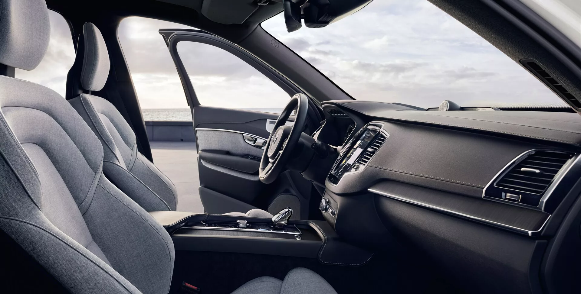 Tento obrázek ukazuje interiér Volvo XC90 druhé generace z roku 2015 z pohledu řidiče. Mírně zvýšený úhel nabízí detailní pohled na přední sedadla a palubní desku, která je vybavena levostranným řidičovým sedadlem čalouněným texturovanou látkou s viditelnou opěrkou hlavy a bočními podpěrami. Multifunkční volant má několik ovládacích tlačítek a elegantní palubní deska obsahuje symetrické vzduchové větráky a částečně viditelný centrální infotainmentový displej. Středová konzola ukazuje řadicí páku automatické převodovky a další ovládací prvky, které zdůrazňují ergonomický a praktický design.