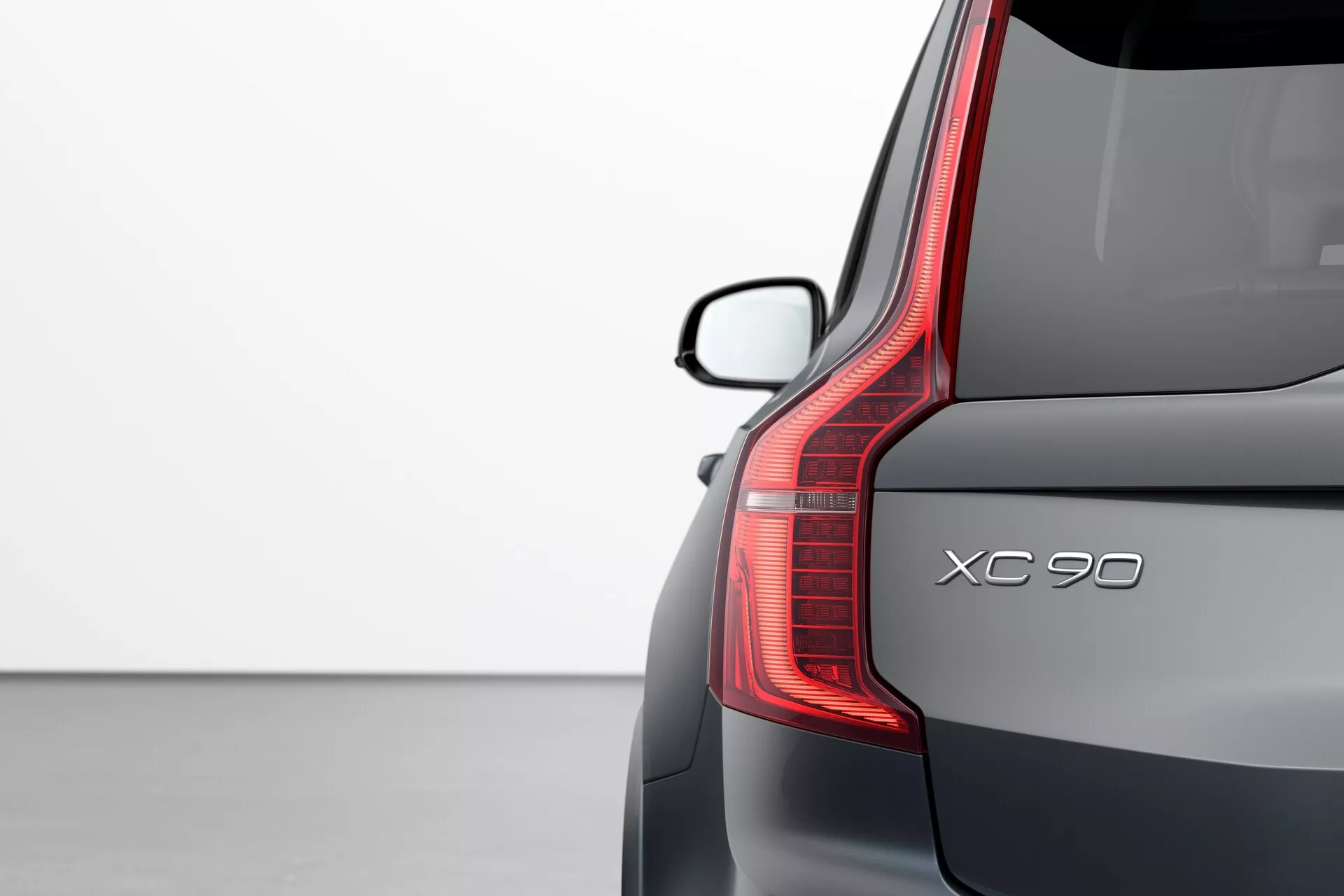 Snímek zadního rohu modelu Volvo XC90 (2. generace) z roku 2015, který zvýrazňuje designové detaily: vertikální zadní světlo LED, název modelu 'XC90', elegantní zadní čelní sklo se spoilerem a prémiovou metalickou barvu na světlém pozadí.