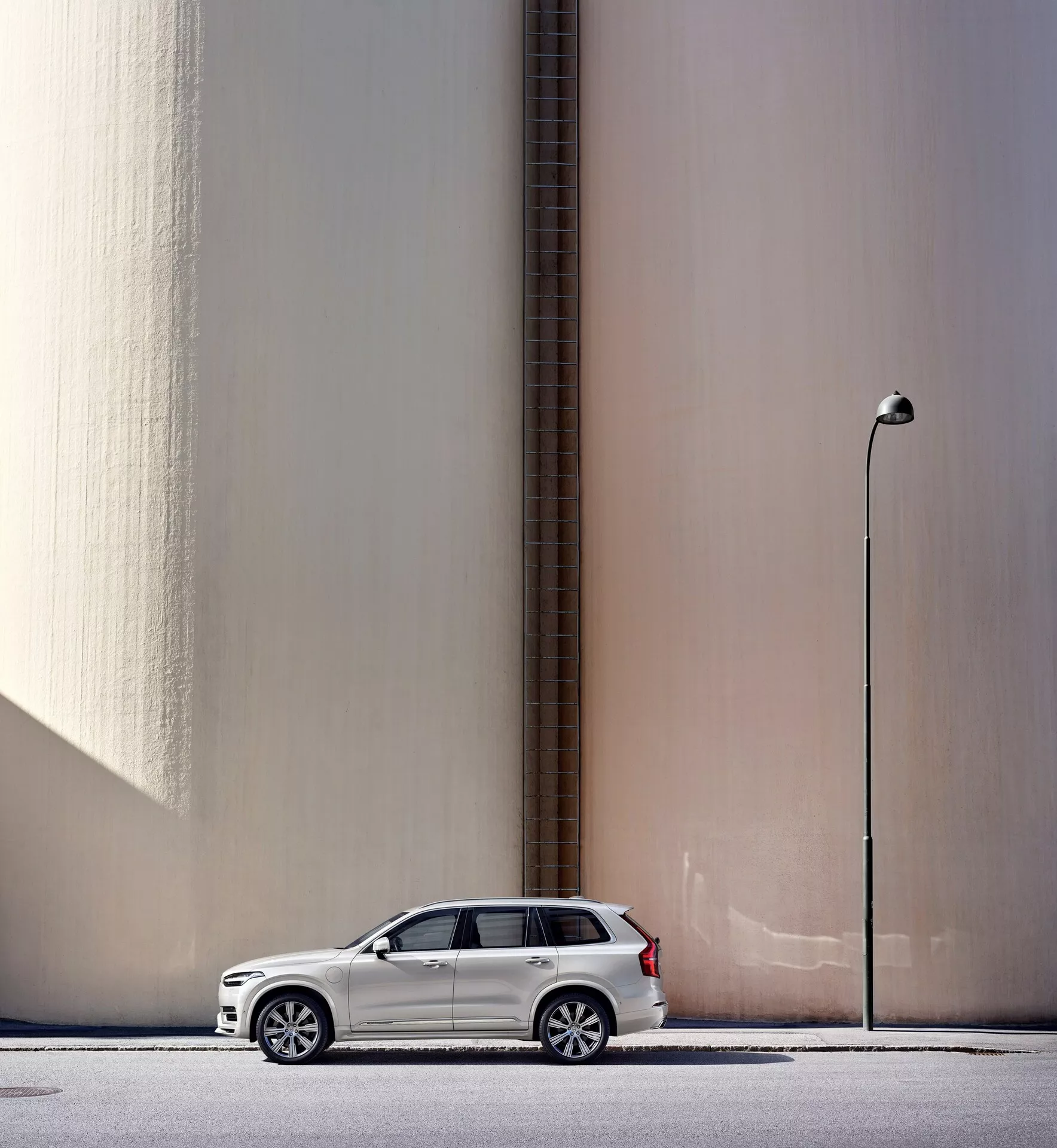 Boční profil bílého SUV Volvo XC90 z roku 2015 zaparkovaného rovnoběžně s vysokou, hladkou béžovou zdí s vertikálním potrubím, na pozadí městské scenérie se sloupem pouliční lampy. Auto, umístěné vlevo v rámu s rozloženými bočními zrcátky, vrhá mírný stín naznačující přirozené denní světlo. Obrázek také ukazuje moderní prvky designu, jako jsou slitinová kola a LED světlomety, které vyzdvihují klíčové automobily.