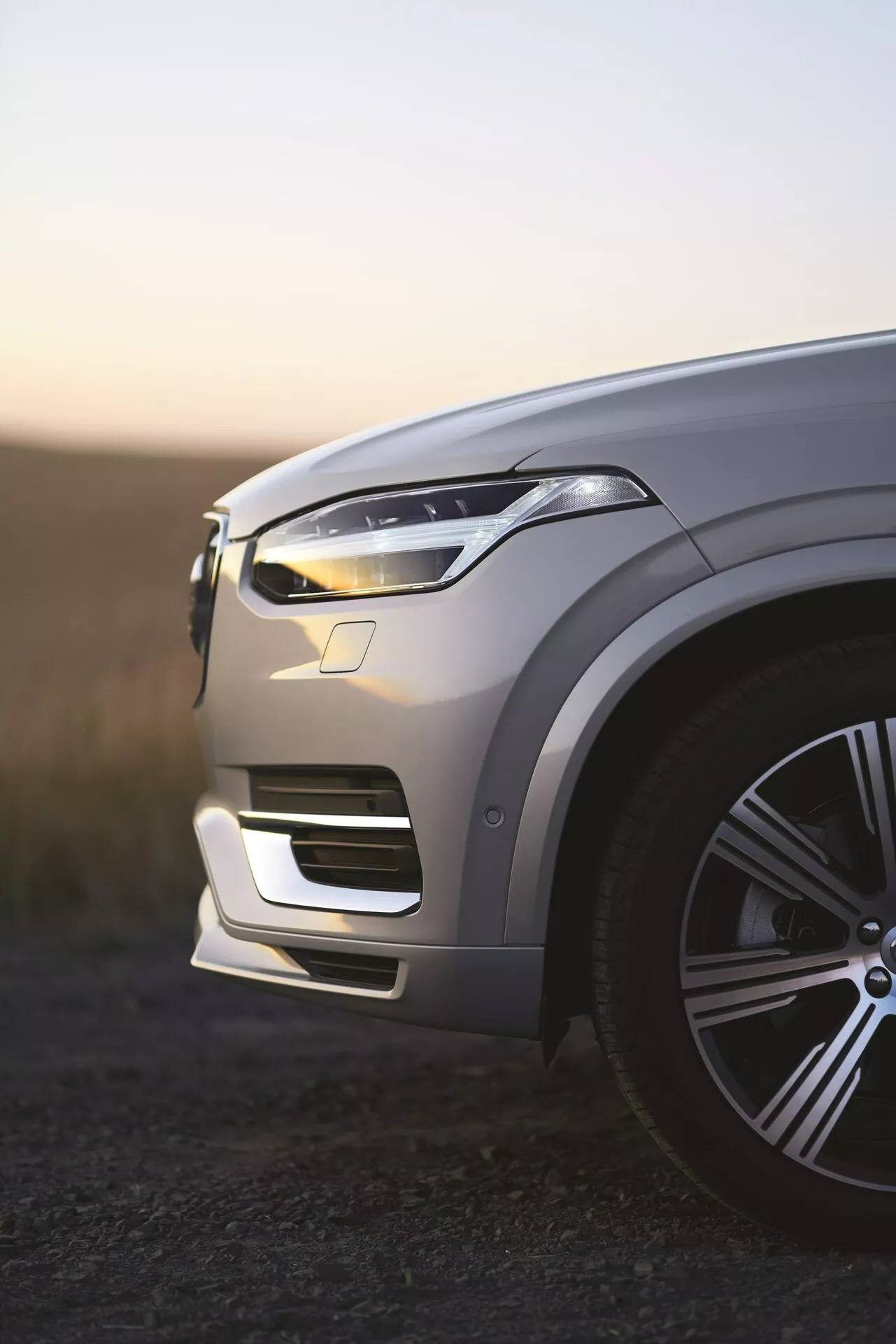 Pohled na levý přední roh luxusního SUV Volvo XC90 2. generace z roku 2015 za úsvitu, osvětlené LED denní světla, kovový lak odrážející okolní světlo, stylové velké litá kola, složité kontury kapoty, elegantní design LED světlometů, integrovaný přední nárazník s nasávači vzduchu, viditelné potenciální senzory asistenta řízení, mělká hloubka ostrosti s rozmazaným pozadím.