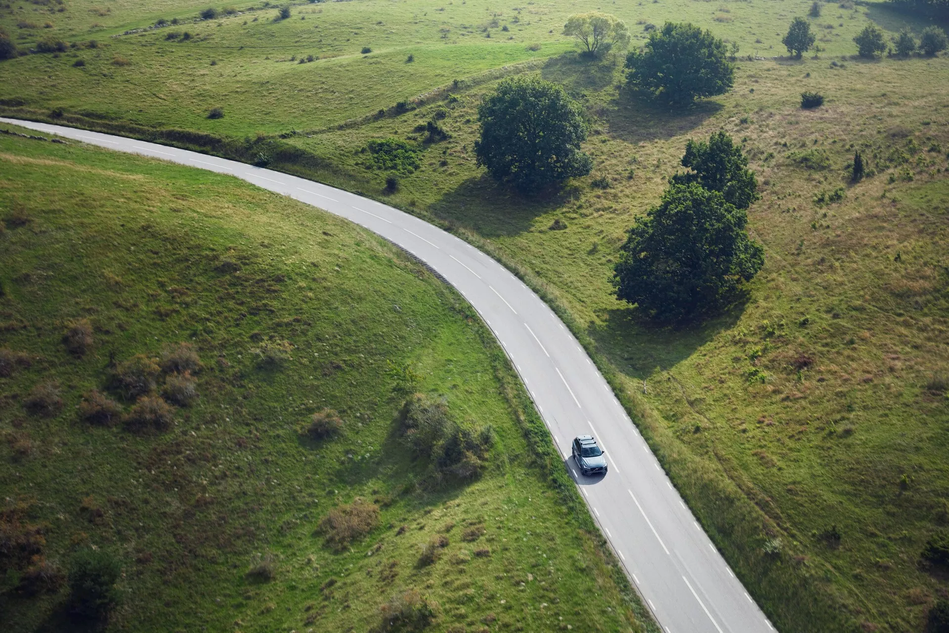 Letecký pohled na stříbrný nebo světle šedý Volvo XC90 z roku 2015, který jede po zakřivené venkovské silnici obklopené travnatými poli s malými stromy a keři. Auto je zachyceno ze shora a je umístěno uprostřed zatáčky vpravo od středu rámu, vrhá stín naznačující polední slunce.