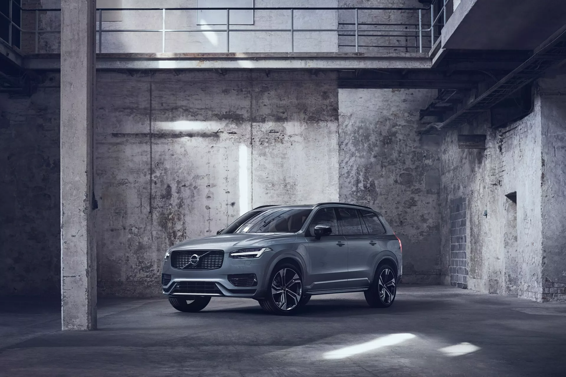 Obrázek kovově zbarveného vozu Volvo XC90 2015 zaparkovaného v temně osvětlené, industriální garáži. Zachycen z pohledu tři čtvrtiny zepředu, předvádějící řidičovu stranu s výrazně zobrazenou přední částí a bočním profilem. Auto má úzké LED světlomety, velkou přední mřížku, možná parkovací senzory a velká hliníková kola s designem s více paprsky. Boční zrcátka jsou aerodynamicky tvarovaná a mohou zahrnovat pokročilé technologické funkce jako indikátory směru a systémy varování mrtvého úhlu. Obrázek zdůrazňuje luxusní, ale praktický design SUV s vysokou světlou výškou a možnými střešními funkcemi, jako je střešní okno nebo panoramatická střecha.