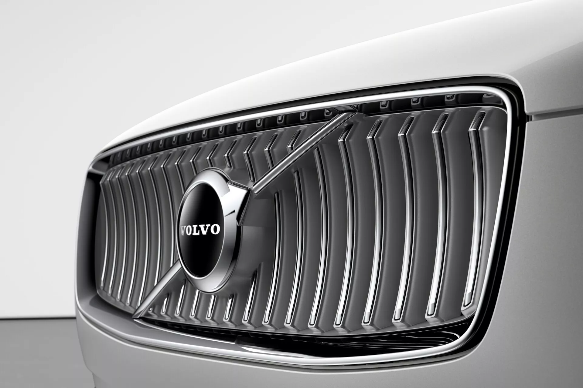 Detailní pohled na přední masku vozu Volvo XC90 2015 (2. generace) z úhlu třech čtvrtin předku, s výrazným znakem Volvo umístěným ve středu svislých chromovaných lamel, pod jemným osvětlením zvýrazňujícím světlou barvu vozu, pravděpodobně bílou nebo velmi světle šedou, a elegantní moderní konkávní design masky.