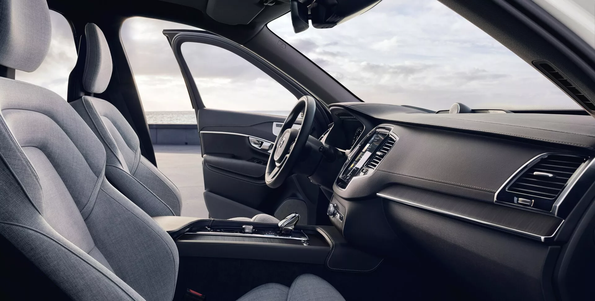 Pohled na interiér vozu Volvo XC90 2015 (2. generace) z perspektivy předního spolujezdce, který ukazuje ergonomické sedadlo řidiče čalouněné tmavou prémiovou textilií nebo syntetickou kůží, multifunkční volant s ovládacími prvky, elegantní palubní desku s vypnutým dotykovým infotainment systémem, integrované vzduchové výdechy, minimalistickými ovládacími prvky klimatizace, středovou konzoli s úložným prostorem, držáky na nápoje a řadicí pákou, vše zvýrazněné jednotným tmavým barevným schématem osvětleným přirozeným denním světlem.