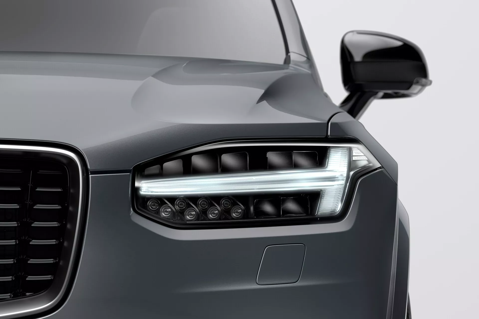 Zblízka pohled na levý přední roh Volvo XC90 z roku 2015 (2. generace) zobrazující moderní LED světlomety s úhlovými denními světly, částečný pohled na velkou mřížku s vertikálním vzorem, lesklý tmavě šedý metalický lak a parkovací senzor na nárazníku, pořízený z nízkého úhlu zprava doleva.