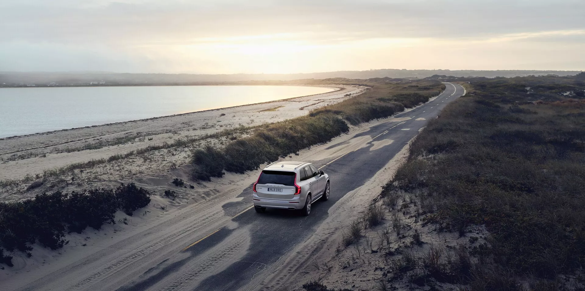 Letecký snímek bílého nebo světle zbarveného vozu Volvo XC90 2015 (2. generace) jedoucího po klikaté prašné cestě rovnoběžné s vodním tělesem, pravděpodobně řekou, s pobřežní krajinou napravo. Scéna se odehrává buď za úsvitu, nebo za soumraku s měkkým přirozeným světlem vrhajícím dlouhé stíny, což zvyšuje detaily vozu a jeho okolí. Vůz je umístěn mírně vpravo od středu ve spodní třetině obrazu, přičemž je vidět jeho zadní část včetně zadních dveří a zadního skla. Obrázek zdůrazňuje pocit dobrodružství a cestování v malebném přírodním prostředí.