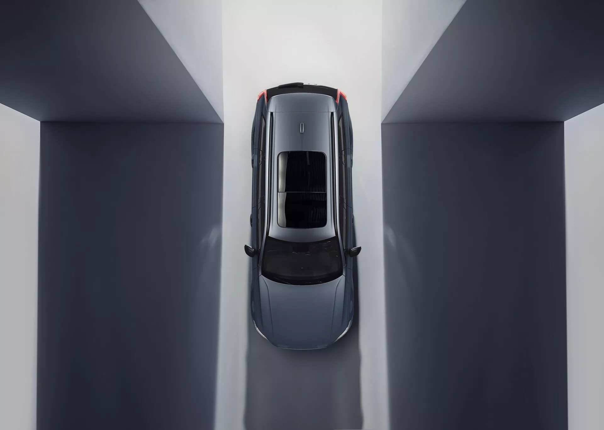Letecký pohled na Volvo XC90 2015 (2. generace) zobrazující symetrickou kompozici a odrazy prostředí. Centrální umístění v neutrálně tónovaném vnitřním prostředí zvýrazňuje střechu vozu se střešním oknem nebo panoramatickou skleněnou střechou. Viditelné části zahrnují kapotu, střechu, kufr a horní polovinu dveří, zdůrazňující elegantní linie a křivky naznačující luxusní design zaměřený na aerodynamiku a prostorné osvětlení interiéru.