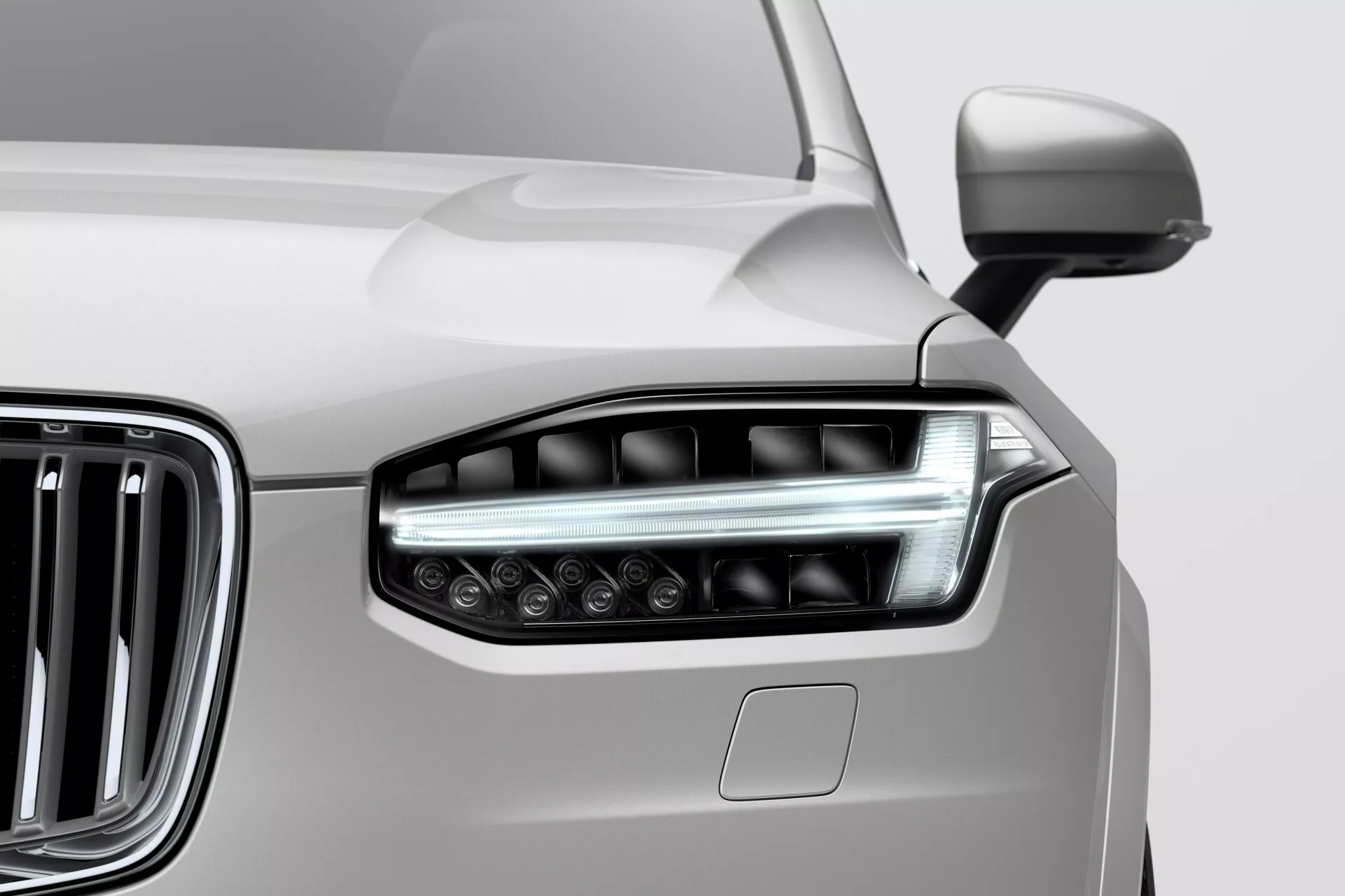 Detailní pohled na přední levé světlomet a část mřížky chladiče z vozu Volvo XC90, rok 2015 (druhá generace). Elegantní světlomet obsahuje protáhlé, pokročilé LED/HID osvětlení s jasným krytem a má hranatý, aerodynamický design. Přilehlá chromová mřížka s vertikálními lamelami zdůrazňuje moderní, luxusní vzhled vozu. Karosérie, pravděpodobně v neutrální barvě jako je stříbrná, zobrazuje plynulý přechod od světlometu. Částečně je také vidět boční zrcátko. Snímek zdůrazňuje moderní styl a pokročilou osvětlovací technologii vozidla.