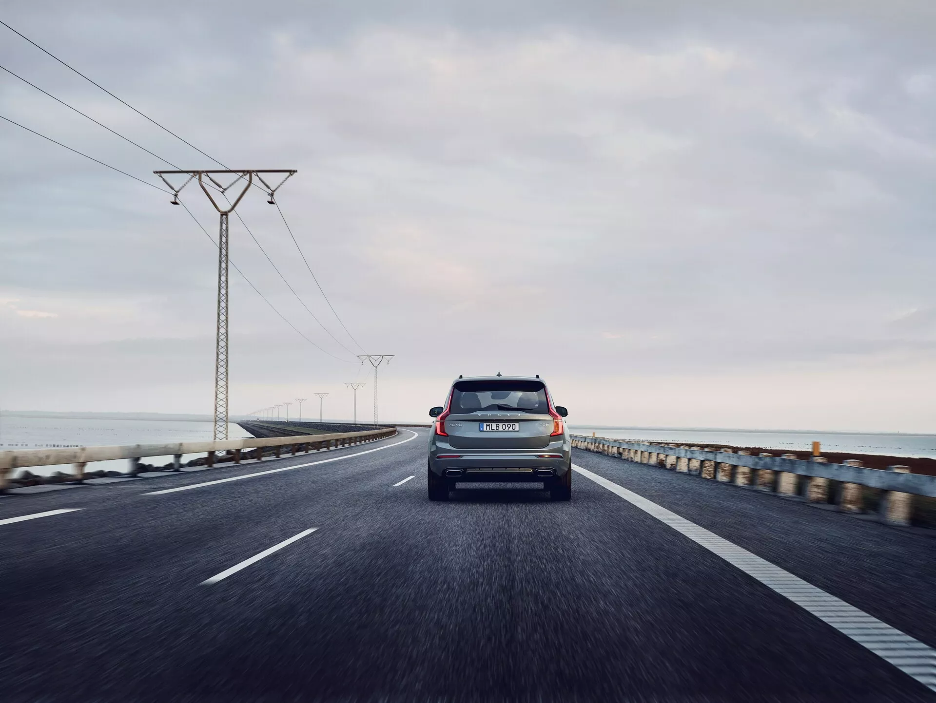 Pohled na zadní část Volva XC90 z roku 2015 (2. generace) cestujícího po dvouproudé přímořské silnici. Fotografie je pořízena z nízkého úhlu, centrální perspektivy, zachycující zavazadlový prostor, zadní sklo, zadní světla, zadní nárazník a označení výrobce a modelu. Vozidlo je hlavním zaměřením, zatímco mírně rozmazaný horizont oceánu a elektrické věže ustupující do dálky pod šedou, zataženou oblohou. RZ vozidla je viditelná a osvětlení naznačuje brzké ráno nebo pozdní odpoledne s rozptýleným světlem.