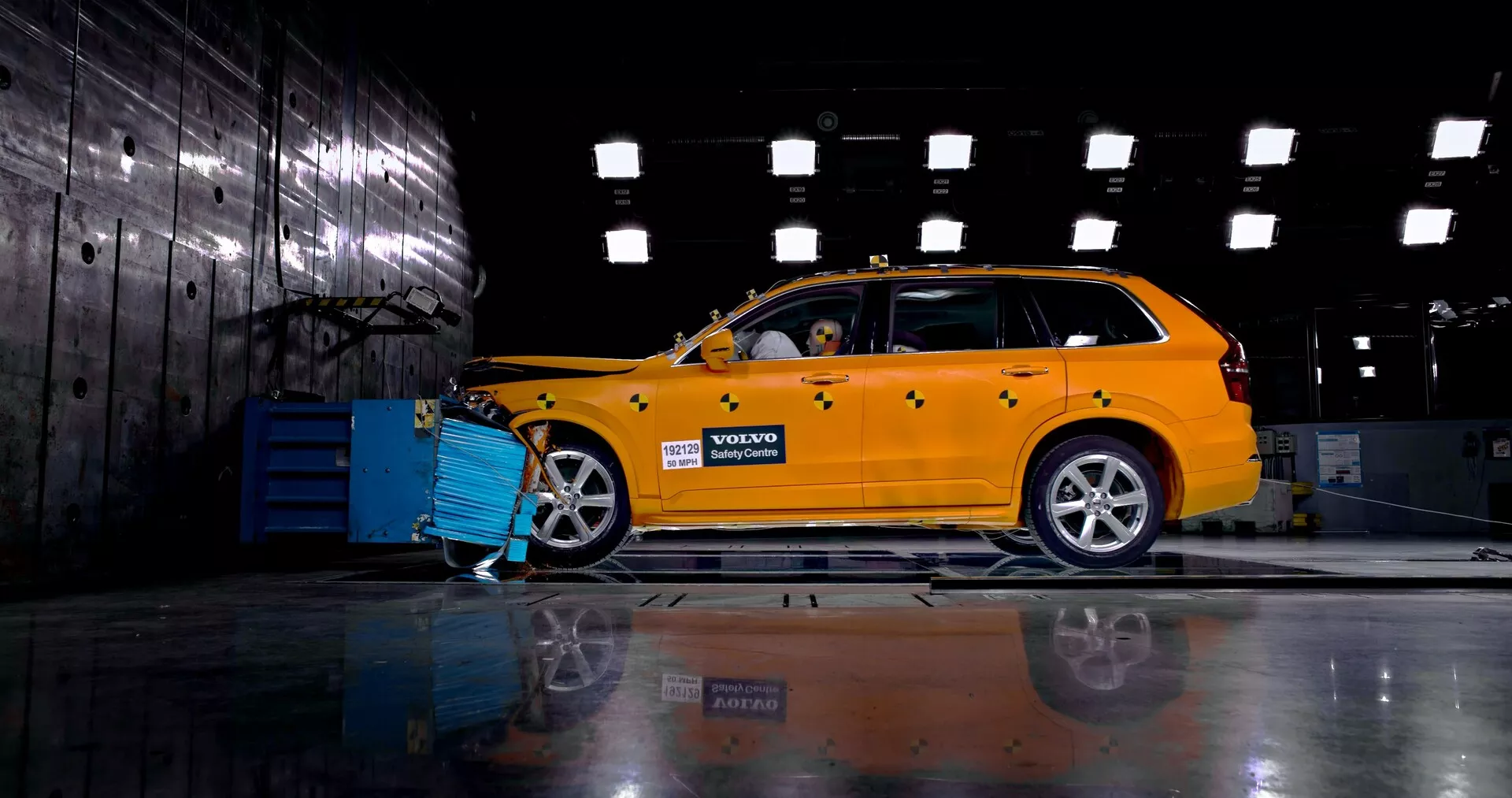 Boční profilový pohled na jasně žlutý 2015 Volvo XC90 (2. generace) v průmyslovém prostředí, který ukazuje jeho design kombi s viditelnou přední mřížkou, světlomety a slitinovými koly. Auto je připojeno k modrému diagnostickému nebo nabíjecímu zařízení a figurína uvnitř naznačuje bezpečnostní nebo nárazový test. Na leštěné podlaze se odráží podvozek a kola na opačné straně.