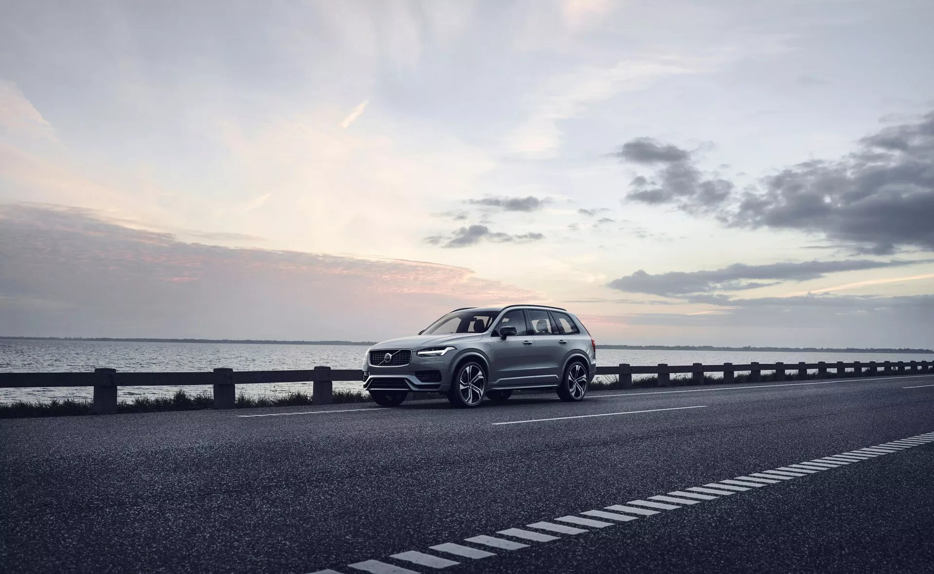 Moderní SUV, konkrétně Volvo XC90 z roku 2015 (2. generace), zaparkované na pravé straně dvouproudé silnice. Vozidlo je zachyceno z tříčtvrtečního předního pohledu, který ukazuje přední mřížku chladiče, světlomety, kapotu a blatník na straně řidiče. Úhel také odhaluje část profilu vozu, včetně kol, dveří řidiče a části zadní části. Kola jsou mírně otočená doprava, s pravděpodobně sportovnějším designem, jako je výrazný nárazník a větší nasávací otvory, což naznačuje dynamický balíček. Fotografie, pořízená pravděpodobně za soumraku s měkkým, rozptýleným světlem pod mírně zataženou oblohou, zvýrazňuje klidné prostředí. Dobře definovaná karoserie s viditelnými liniemi naznačuje luxusní model. Mezi technologické prvky, které lze z fotografie vyvodit, patří LED světlomety, pokročilé designy kol, sofistikovaná přední mřížka s možnými senzory nebo kamerami a aerodynamické prvky. Tento kvalitní obrázek by mohl být vhodný pro reklamy, zdůrazňující vůz jako luxusní volbu životního stylu.