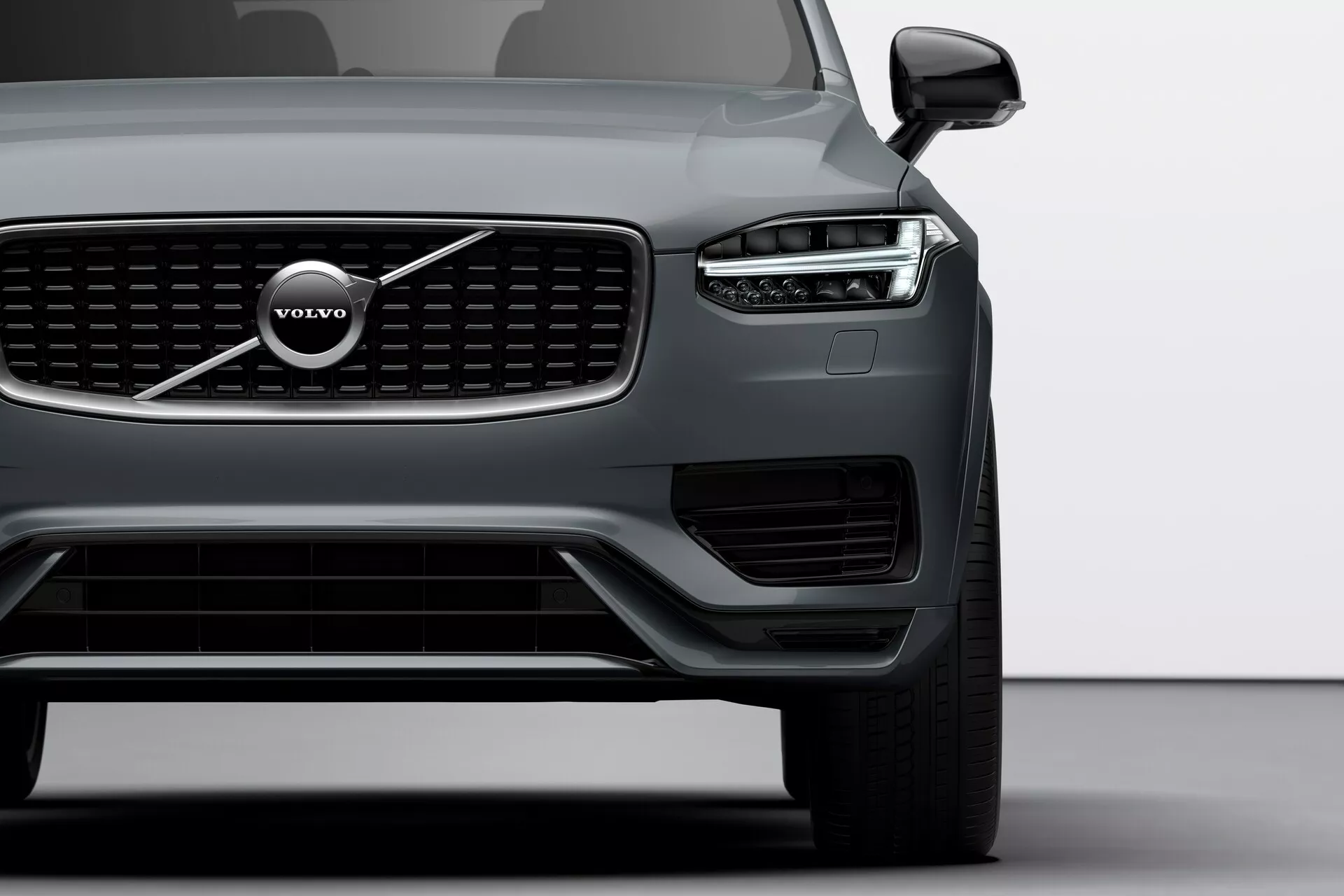 Pohled z levého předního čtvrtletníku na Volvo XC90 z roku 2015 (druhá generace), z nízkého úhlu, který ukazuje ostrá, úhlová přední světla s charakteristickými LED denními světly ve tvaru Thorova kladiva, výraznou přední mřížku s vertikálními lamelami a emblémem Volvo, širokým dolním přívodem vzduchu s dodatečným osvětlením/senzory, aerodynamickými obrysy kapoty, elegantním vnějším zrcátkem s integrovaným směrovým ukazatelem, částečně viditelným velkým vícelamelovým litým kolem a matným povrchem karoserie, zdůrazňující moderní design a pokročilé technologie vozidla.