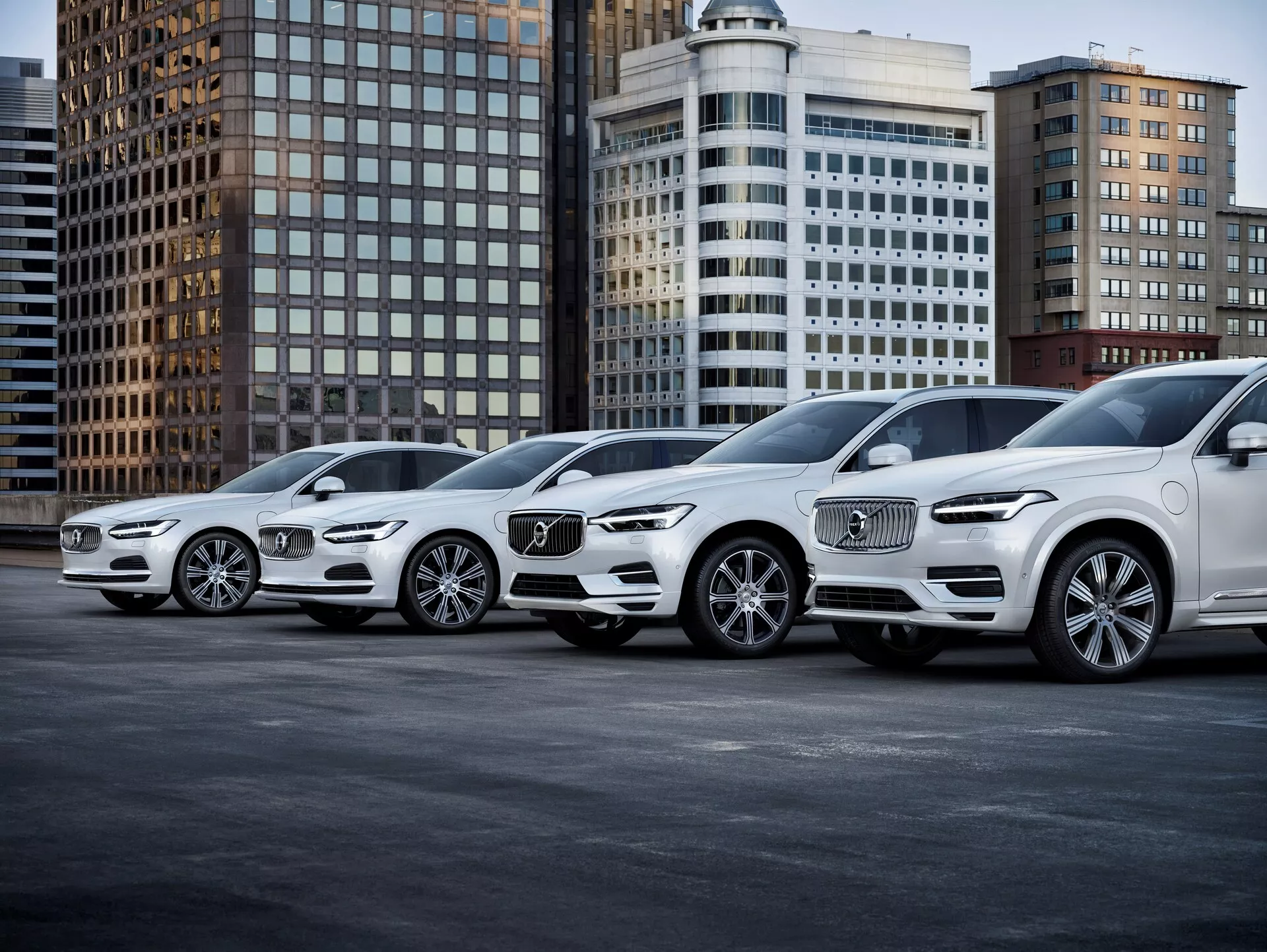 Obrázek zobrazuje řadu čtyř luxusních SUV Volvo XC90 druhé generace z roku 2015, které jsou nakloněny s předním profilem směřujícím doprava. Umístěny diagonálně, perspektiva zvýrazňuje jak přední, tak boční pohledy vozidel. Auta mají výrazné přední mřížky s rozpoznatelným logem Volvo, složité LED světlomety, designy slitinových kol a kontury na kapotě. Fotografie, pořízená z nízkého úhlu blízko země, zdůrazňuje velikost a postoj SUV, zasazených do městského pozadí s kancelářskými budovami. Osvětlení je měkké, což naznačuje zatažený den nebo dobře osvětlené městské prostředí, jasně ukazující kola, světlomety, mlhová světla a designové linie. Nejbližší vozidlo zobrazuje pokročilé technologie osvětlení a přední senzory mřížky, pravděpodobně spojené s autonomním řízením nebo asistencí při parkování. Jak vozidla ustupují, jejich identický styl naznačuje, že patří do stejné rodiny vozidel, což činí tento obrázek ideálním pro kategorie jako Automobilismus, Luxusní vozidla, Pokročilé automobilové technologie a Městský životní styl.