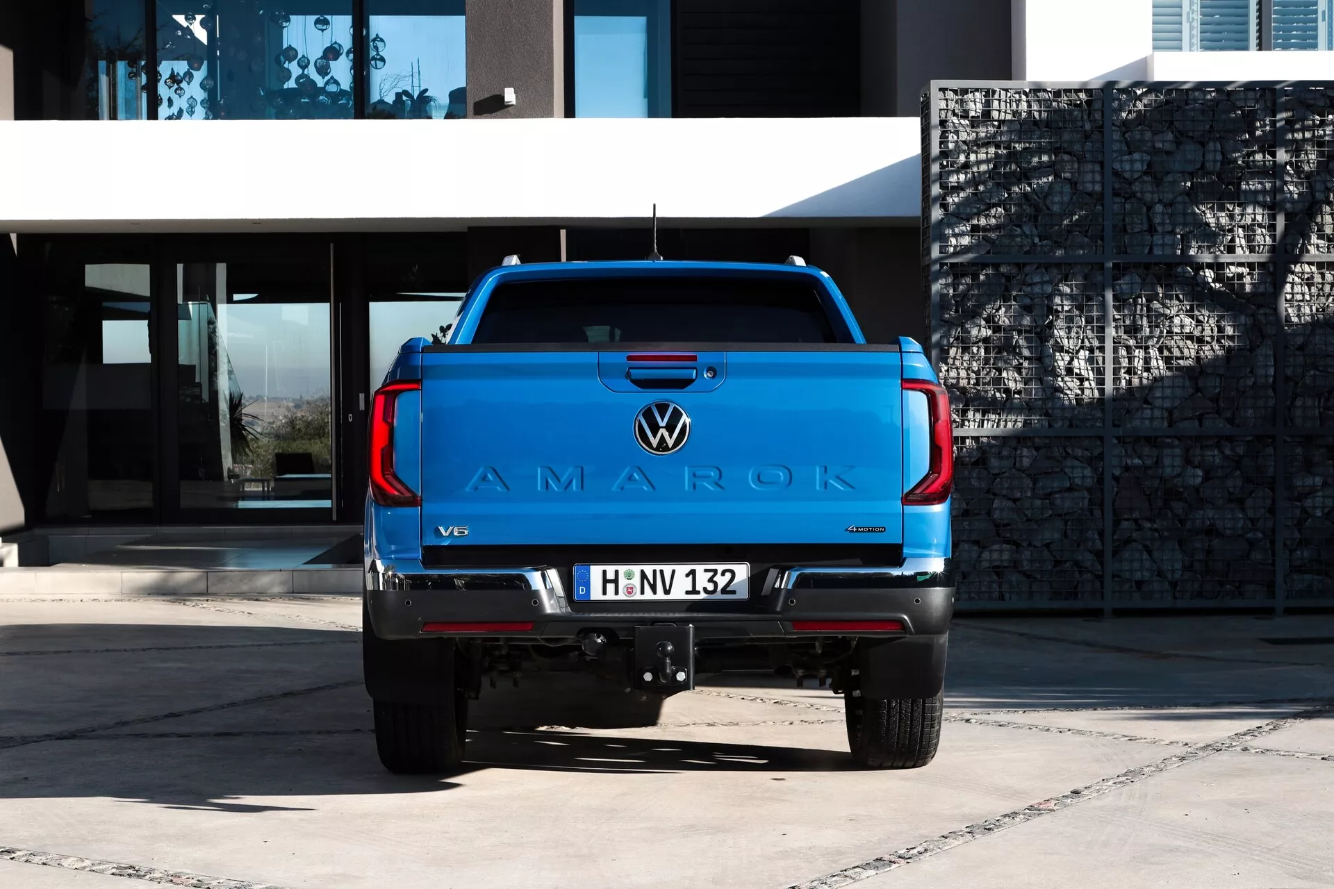 Pohled zezadu na pickup Volkswagen Amarok 2023 s živou modrou barvou na dlážděném povrchu, zobrazující velké logo 'AMAROK' na víku nákladového prostoru, znak VW, odznak motoru V6, moderní zadní světla, tažné zařízení, parkovací senzory, evropskou poznávací značku a dvojitý výfukový systém na pozadí moderní budovy v přirozeném denním světle.