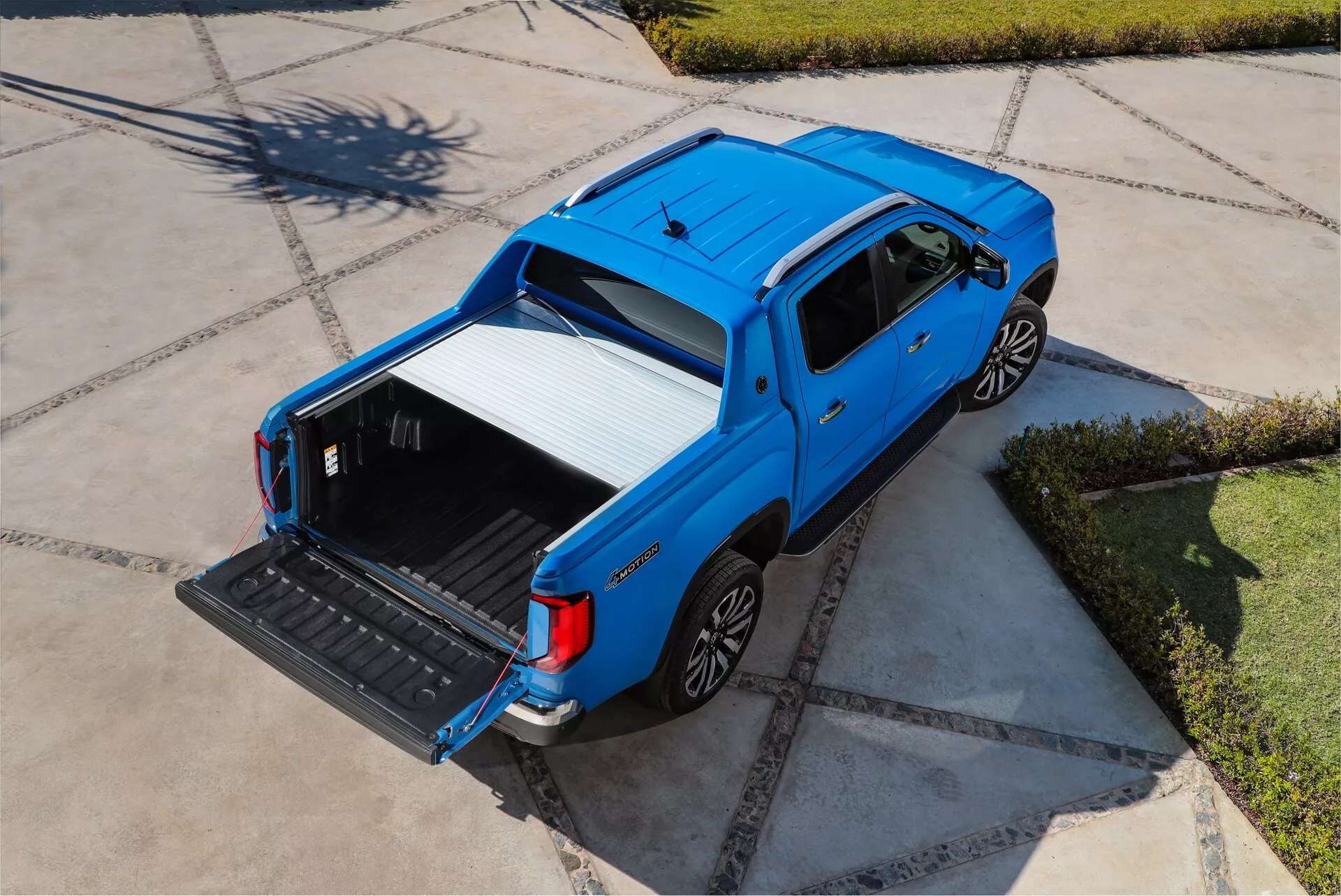 Letecký snímek modrého pickup trucku VW-Nutzfahrzeuge Amarok 2. generace z roku 2023, který zobrazuje jeho horní a boční profil z vysokého úhlu. Vozidlo je zaparkováno na charakteristicky vydlážděné příjezdové cestě, zaplavené slunečním světlem. Záběr z mírně zadního šikmého pohledu zdůrazňuje otevřený nákladní prostor s dole sníženým zadním víkem, který odhaluje odolné, ochranné obložení pro náklad. Elegantní, současný čtyřdveřový design posádky je zvýrazněn bočními zrcátky a klikami dveří v barvě karoserie. Kola mají vícepaprskový design z lehké slitiny, což zlepšuje jak estetiku, tak případný výkon.