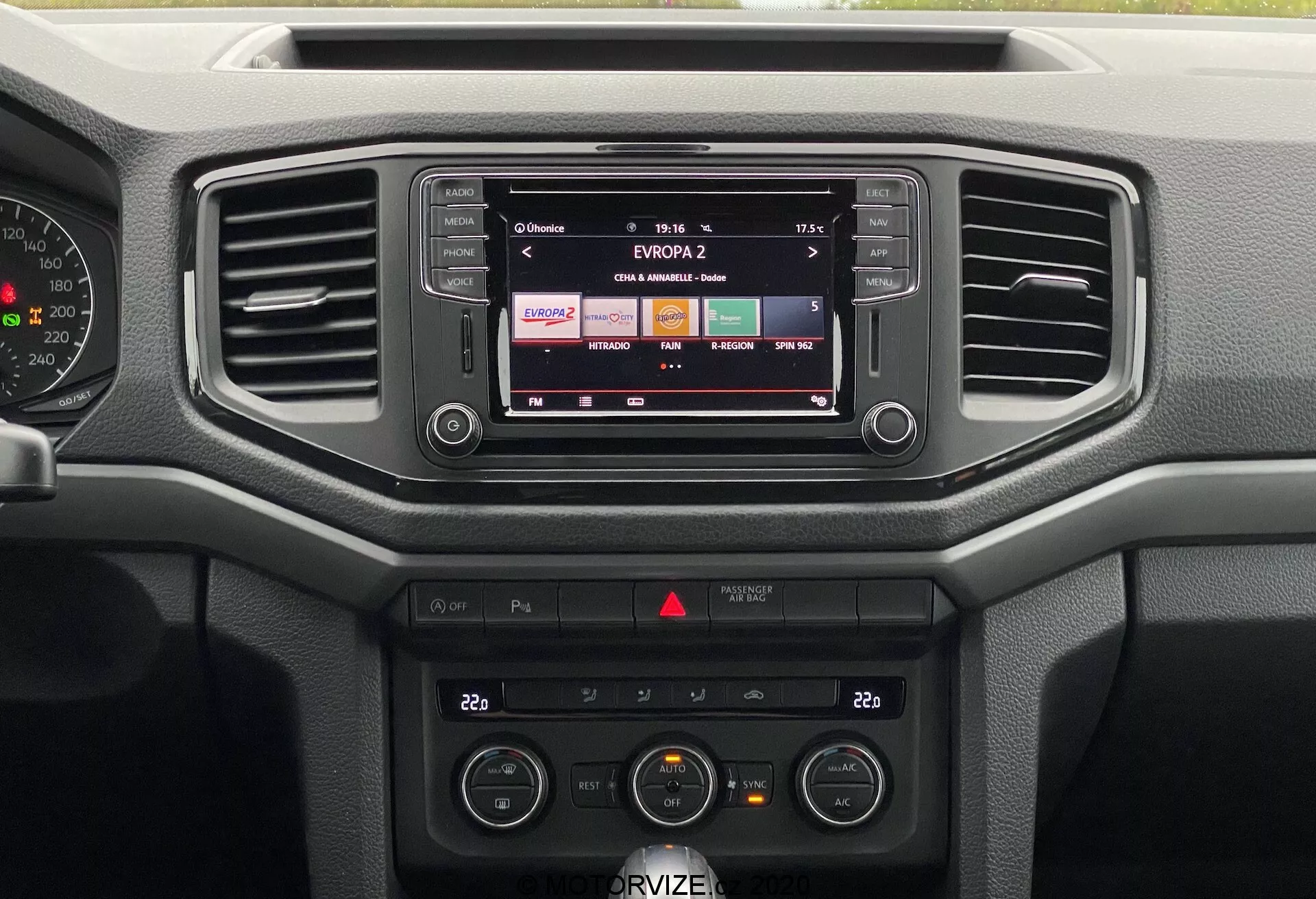 Obrázek představuje centrální palubní desku vozu VW Nutzfahrzeuge Amarok 2016. Z předního pohledu je vidět infotainment systému zobrazujícího rozhlasovou stanici, dva horizontální vzduchové větrací otvory a různé ovládací prvky. Zvukový systém a ovládání klimatizace, včetně nastavení teploty a ventilátoru, jsou jasně viditelné. Palubní deska upřednostňuje funkčnost s lehce použitelnými tlačítky a volicími kolečky, využívajícími především tvrdých plastových materiálů, vhodných pro každodenní použití spíše než luxus.