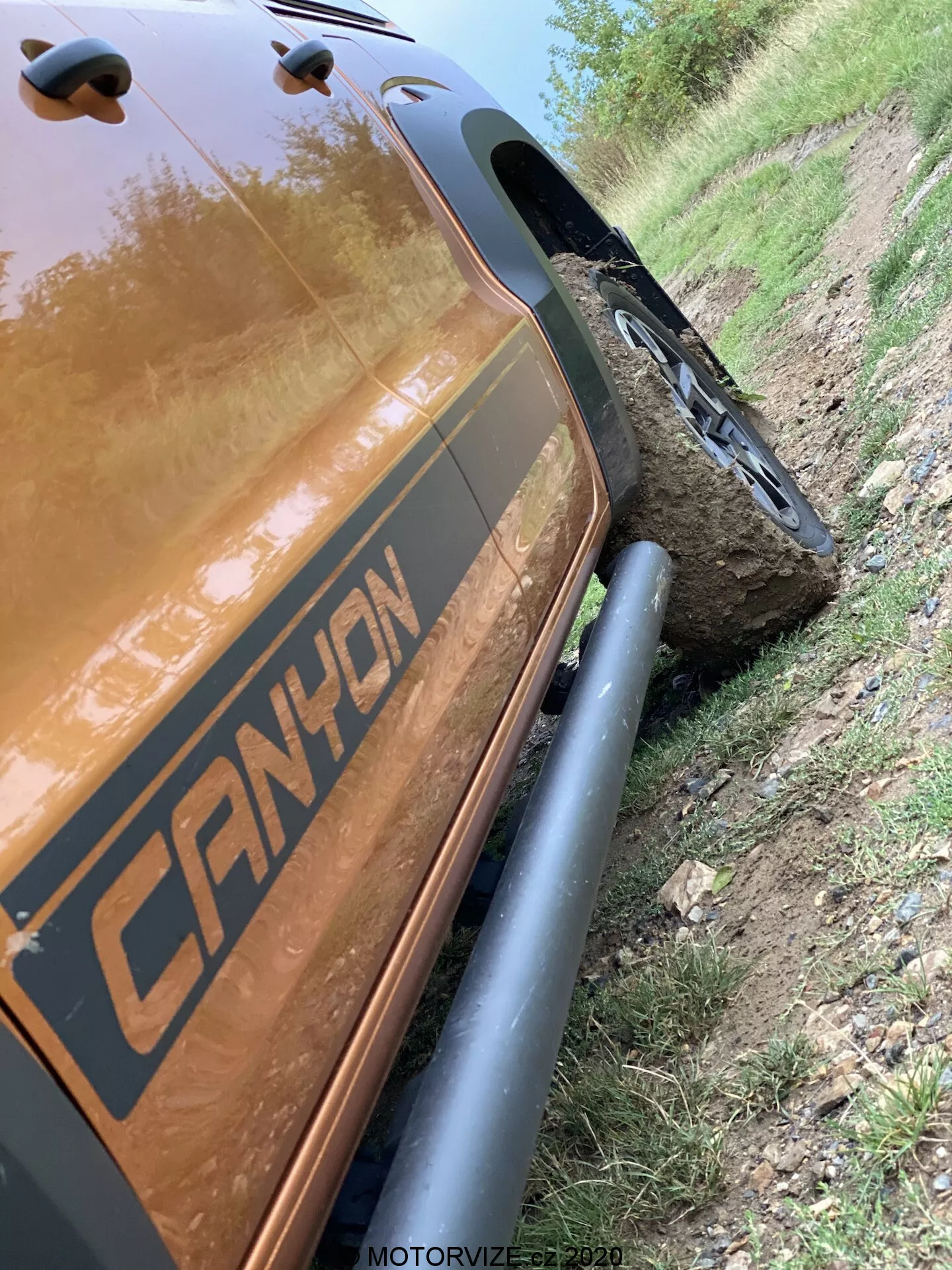 Tento sklonený 45 stupňový obraz zobrazuje zadní část na straně spolujezdce VW-Nutzfahrzeuge Amarok 2016. Zaměřuje se na detailní snímek od zadního blatníku po zadní úložný prostor, zdůrazňuje robustní ochranné kryty blatníků a robustní terénní pneumatiky. Vozidlo má grafiku s nápisem 'CANYON' a je postaveno vedle hliněné cesty s kolem namontovaným na velkém kameni, což dokazuje jeho terénní schopnosti. Reflexní metalická hnědá nebo měděná barva karoserie je výrazná, zachycuje odlesky světla. Časový razítko 'MOTORVIZE.cz 2020' naznačuje, že byl pořízen pro motoristický časopis.