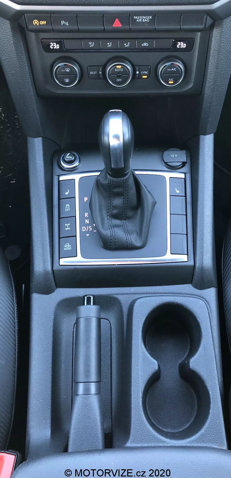 Tento snímek ukazuje středovou konzoli vozu vw-nutzfahrzeuge amarok 2016 z nadhledového úhlu, zhruba z pohledu řidiče nebo předního spolujezdce. Fotografie zachycuje interiér vozu specifický pro řadicí páku, ovládání středové konzole a úložné prostory. Snímek obsahuje ovládací prvky klimatizačního systému, koženě obloženou řadicí páku zasazenou do stříbrného panelu indikujícího automatickou převodovku, ovládání elektronické parkovací brzdy, úložný prostor, dva držáky na nápoje a další úložný prostor směrem k zadním sedadlům. Materiály zahrnují plast, kovové doplňky a kůži, což odráží kombinaci funkčnosti a estetiky.