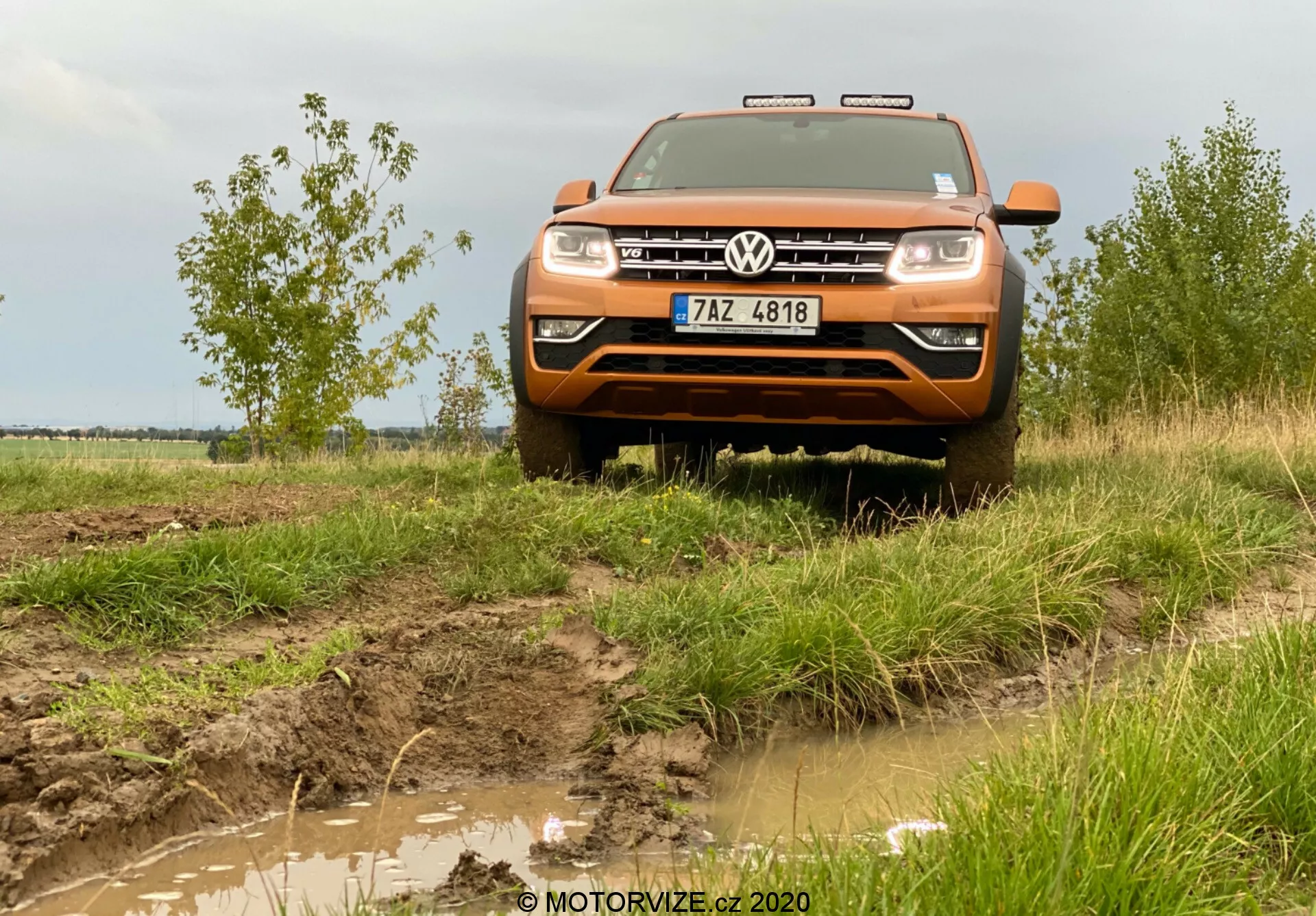 Pohled zepředu pod nízkým úhlem na oranžový Volkswagen Amarok 2016, středně velký pickup, který ukazuje své schopnosti jízdy v terénu. Obrázek zachycuje přední část vozidla, včetně kapoty, mřížky, nárazníku, částí blatníků, světlometů a loga Volkswagen umístěného uprostřed mřížky. Nad mřížkou jsou vidět chromové akcenty nebo LED denní světla. Bahnitý terén a tráva kolem vozu zdůrazňují jeho terénní povahu, a dodatečná střešní světla a přední ochranný kryt s venkovským pozadím s oblačnou oblohou dokončují dobrodružné prostředí. Obrázek je pořízen ze vzdálenosti přibližně dvou až tří metrů, což poskytuje detailní pohled na přední části vozidla a jeho interakci s prostředím.