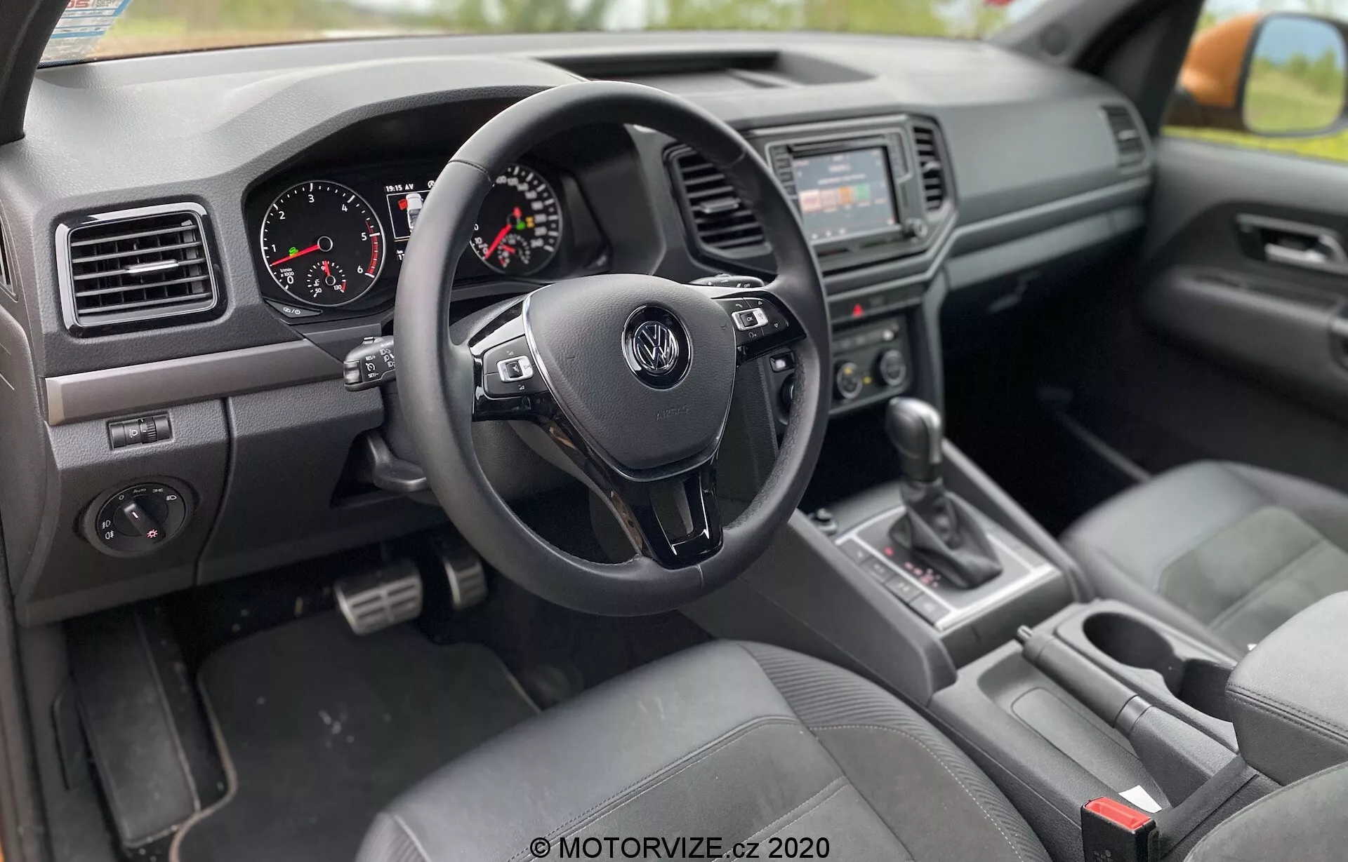 Pohled na interiér VW Nutzfahrzeuge Amarok 2016 z perspektivy spolujezdce, s pravostranným řízením, tříramenným volantem Volkswagen s ovládacími tlačítky, přístrojovým panelem s analogovým rychloměrem a otáčkoměrem, středovou konzolí se zvukovým systémem a infotainment obrazovkou, řadicí pákou v poloze 'P', sedadly potaženými látkou, palubní deskou s ventilačními otvory a texturovanými plasty a ovládacím panelem oken a zrcátek ve dveřích řidiče.