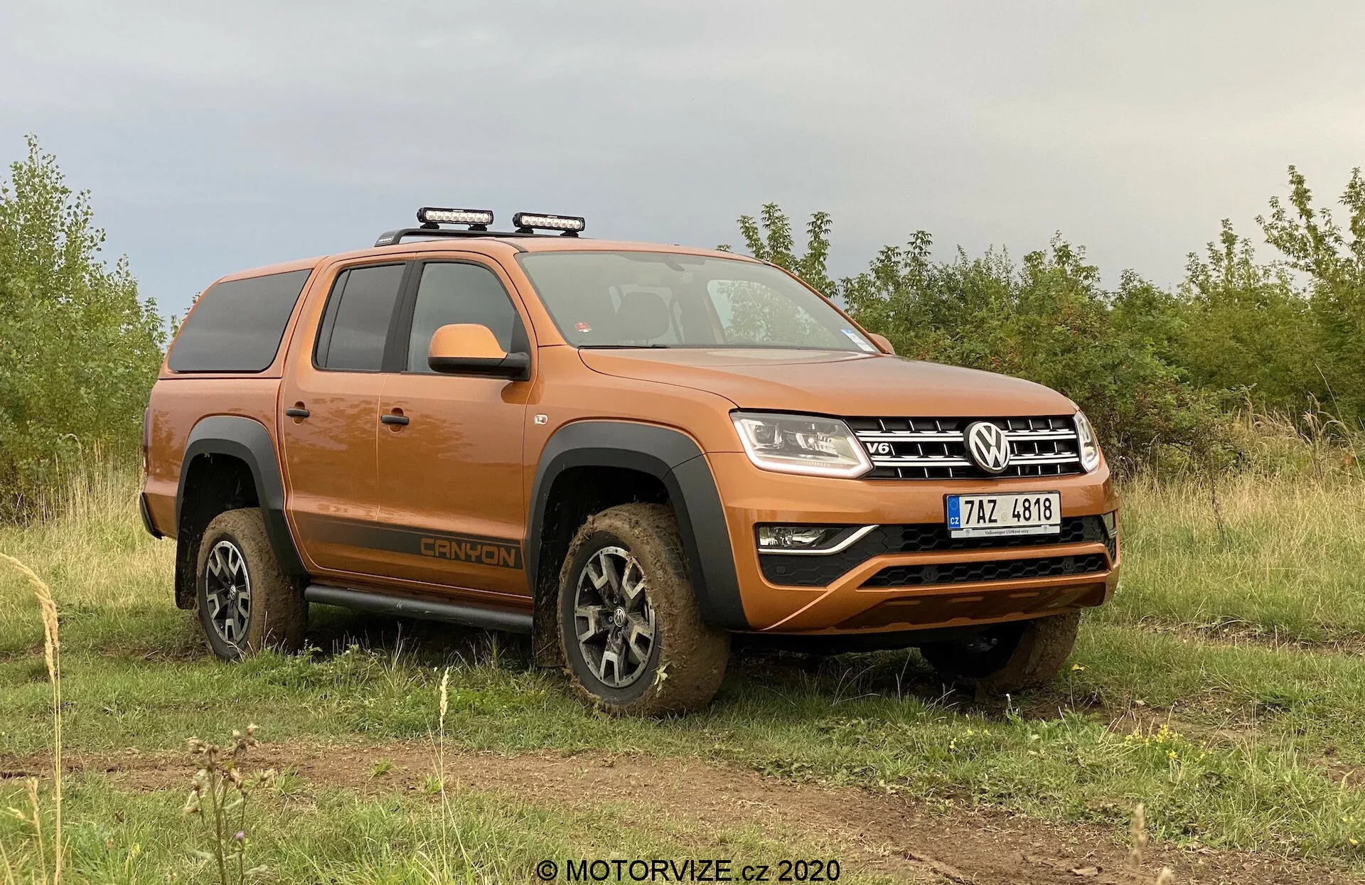 Obrázek zobrazuje oranžový pickup Volkswagen Amarok 2016 zaparkovaný na nerovném travnatém terénu s pozadím oblačné oblohy. Pohled ze strany-přední části se zaměřuje na řidičovu stranu, ukazuje profil vozidla a částečný pohled na přední část. Klíčovými viditelnými prvky jsou střešní nosič, koly pokryté blátem označující nedávnou jízdu v terénu, boční stupátka, konfigurace s dvojitou kabinou, moderní design světel a přední nárazník s mlhovými světly a spodním ochranným krytem. Vozidlo vyzařuje drsný vzhled terénního užitkového vozidla s moderními technologickými prvky.