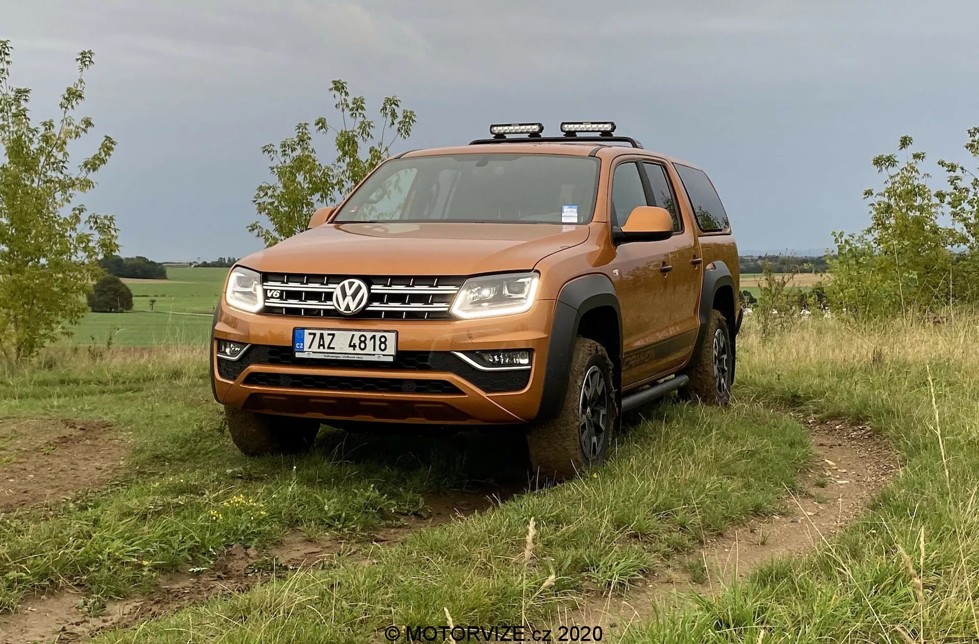 Oranžové SUV Volkswagen, konkrétně model VW Amarok 2016, je zobrazeno na travnatém terénu. Snímek z nízkého až středního úhlu zepředu zdůrazňuje robustní design vhodný pro off-road aktivity. Vozidlo má výraznou přední mřížku s dvěma chromovanými pruhy rámujícími centrální logo Volkswagen, značný nárazník s možným krytem proti skluzu, čiré kompozitní světlomety s potenciálními LED nebo halogenovými žárovkami a rozšířené blatníky s off-road pneumatikami. Na střeše jsou namontována dodatečná světla, což naznačuje větší funkčnost pro noční jízdu. Viditelné detaily zahrnují čelní sklo, střešní ližiny, zpětná zrcátka s integrovanými ukazateli směru a přední registrační značku. Vozidlo vypadá čistě s trochou špíny na spodních stranách a kolech, což odpovídá off-road použití. Pozadí naznačuje venkovské nebo polosoukromé prostředí.